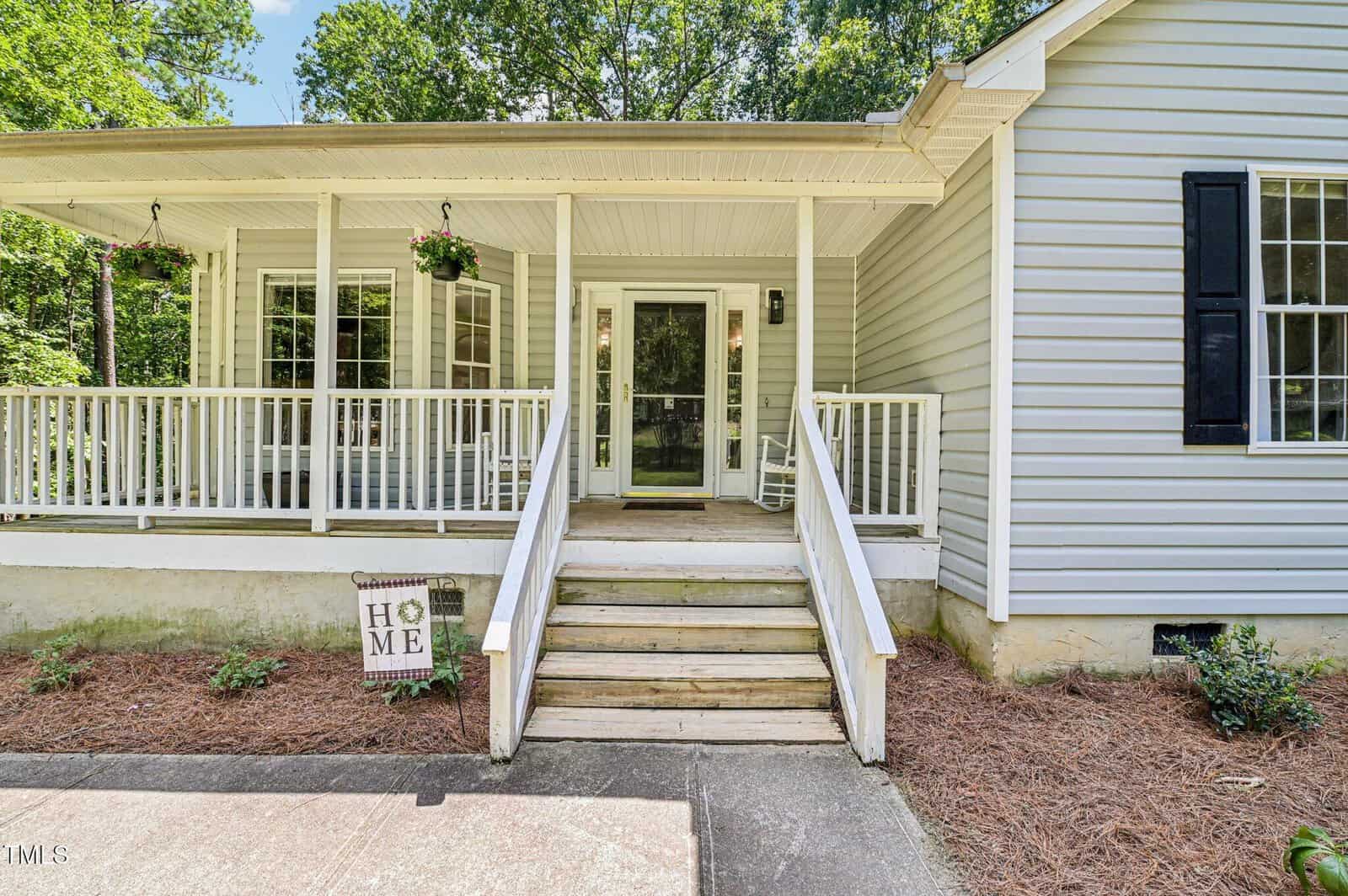 A light gray house with a covered front porch, black shutters, steps leading up, a rocking chair, hanging plants, and a “Home” sign by the walkway. Tall trees and greenery surround the house in the background.