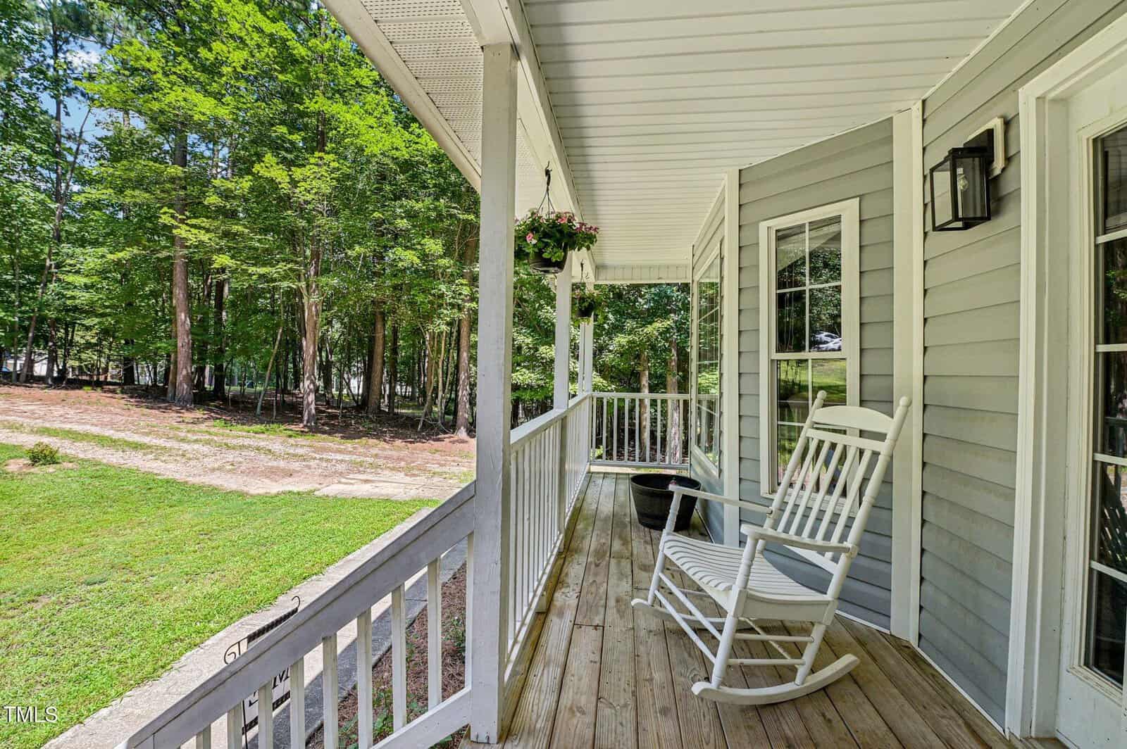 A covered wooden front porch with white railings features a white rocking chair and a hanging plant. The porch overlooks a grassy yard with trees in the background.