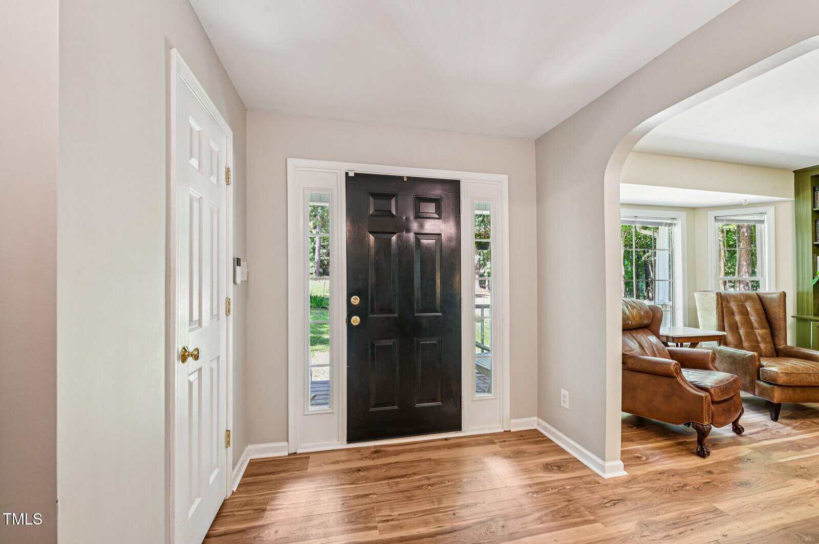 A front entryway with a black door, white trim, and wooden floors. To the right is an open archway leading to a living area with brown leather chairs and large windows. A white closet door is on the left wall.