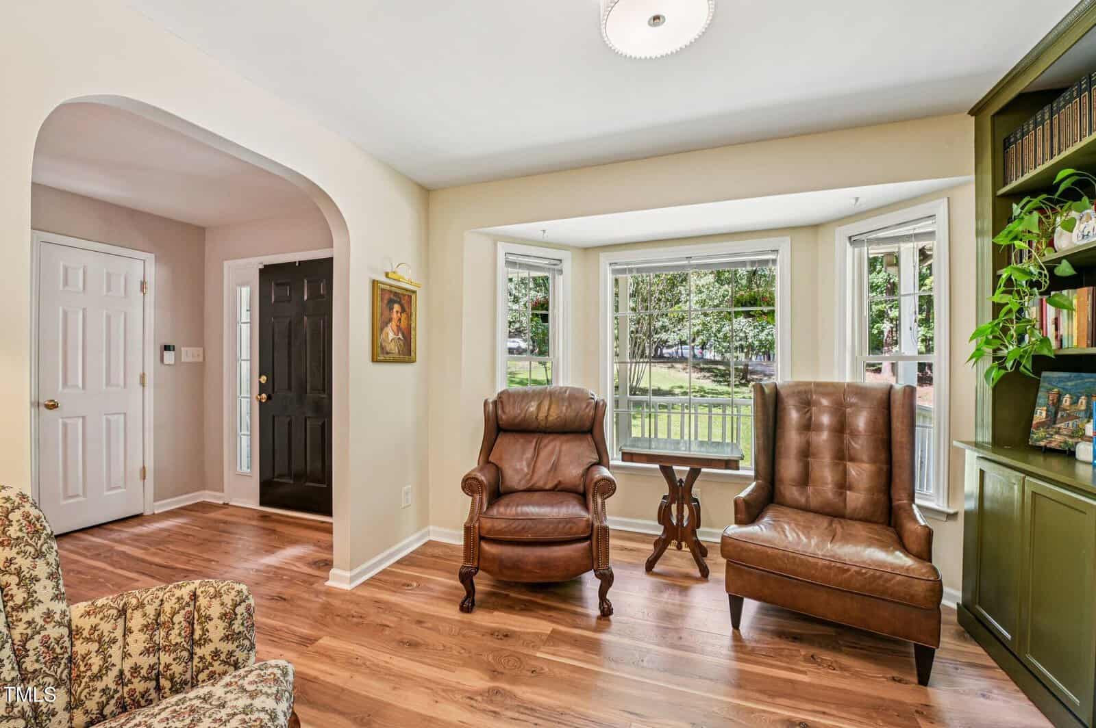 A living room with hardwood floors, two brown leather armchairs near a bay window, a small round table, a floral upholstered chair, green built-in shelves with books and plants, and an arched entryway leading to a foyer.