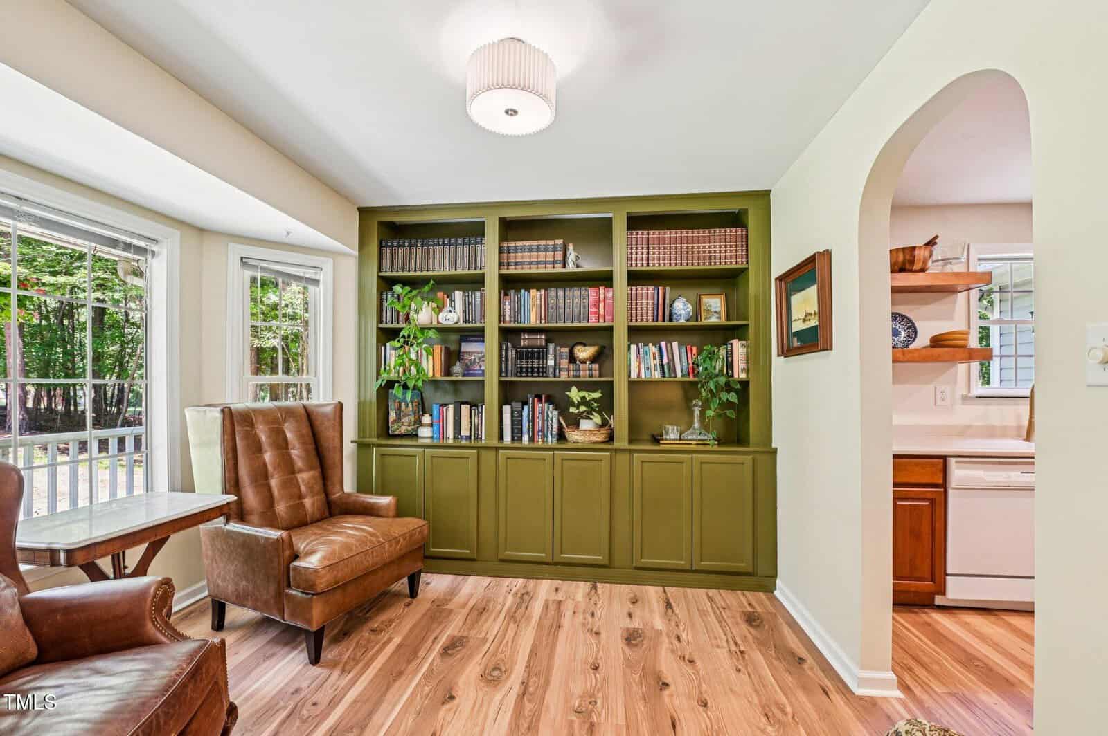 A living room with wood flooring, two brown leather chairs, a built-in green bookcase filled with books and plants, a window with outdoor view, and an arched doorway leading to a white kitchen.