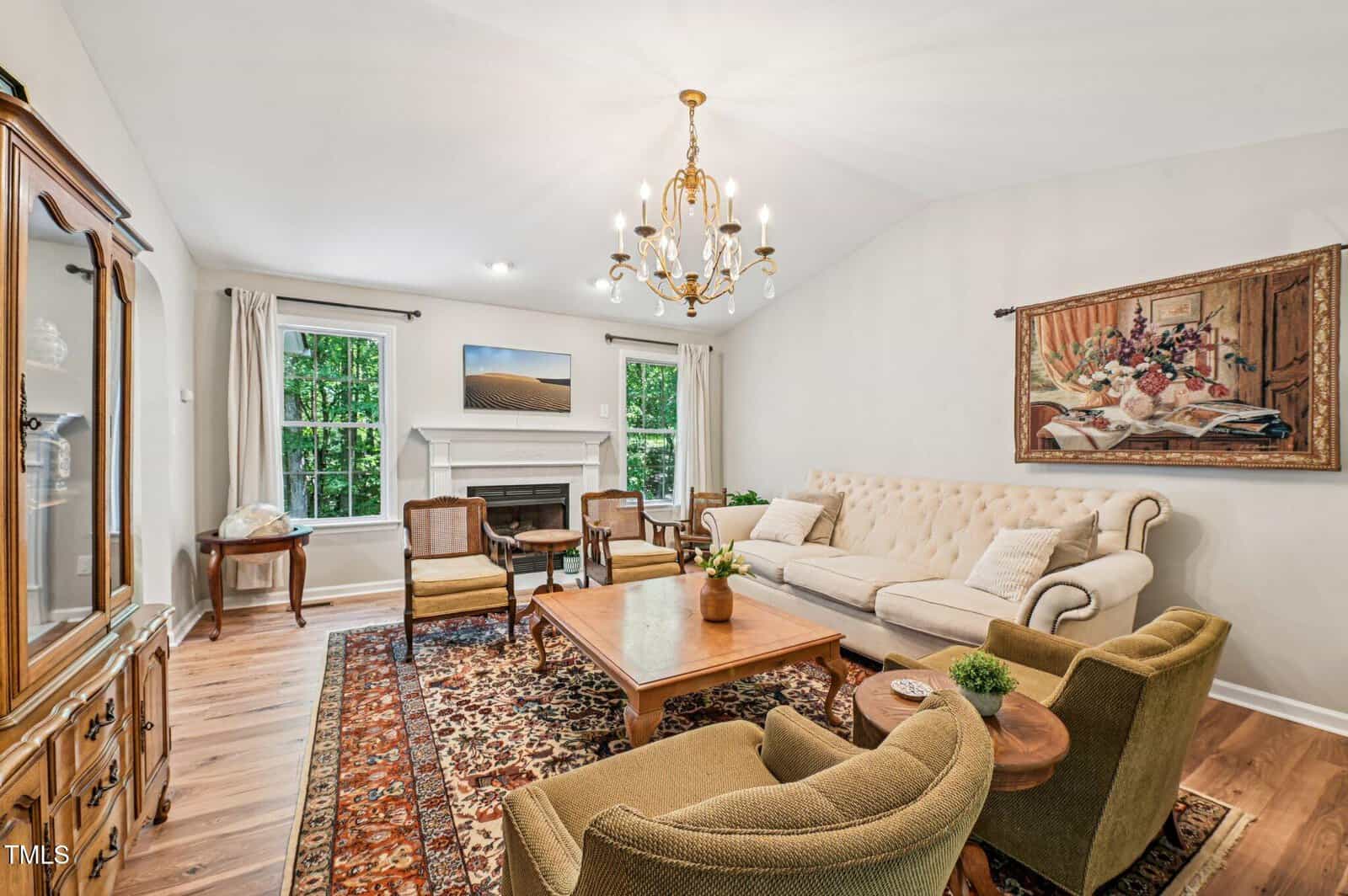 A living room with a beige sofa, two green armchairs, a wooden coffee table, and a patterned rug. There is a fireplace between two windows, a chandelier, and a framed painting on the wall.