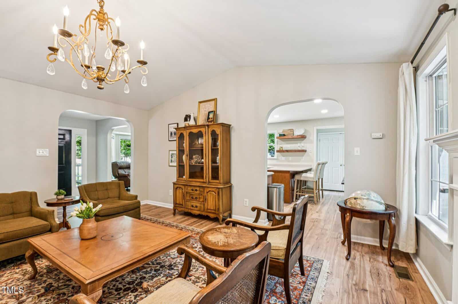 A living room with two armchairs, a wooden coffee table, and a vintage hutch. A globe sits near a window, and a chandelier hangs from the ceiling. The room opens into a kitchen with barstools and white cabinetry.