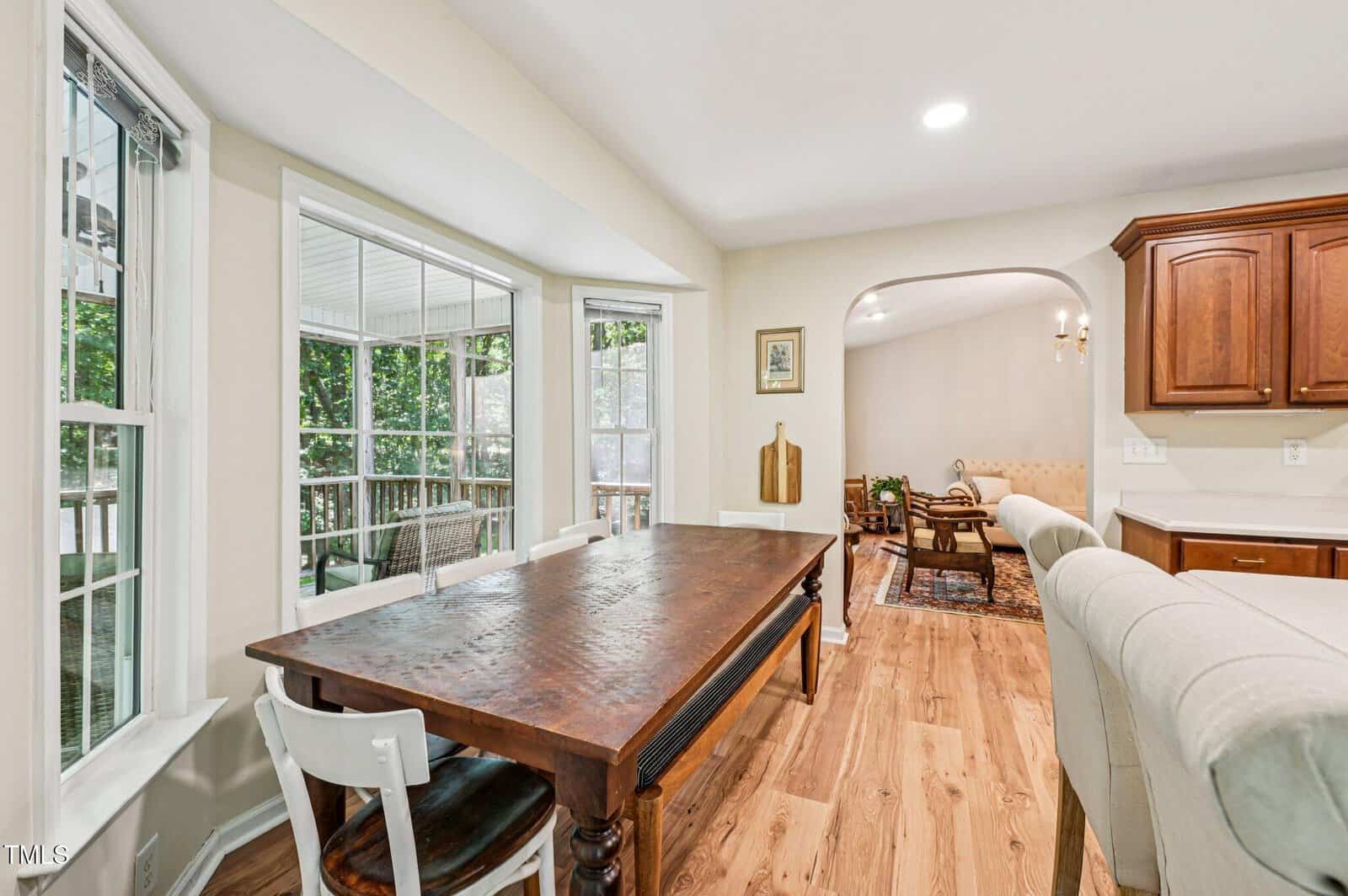 A dining area with a wooden table and mixed chairs sits beside large windows overlooking a porch. The space opens into a living room with wood floors and traditional decor. Kitchen cabinetry is visible to the right.