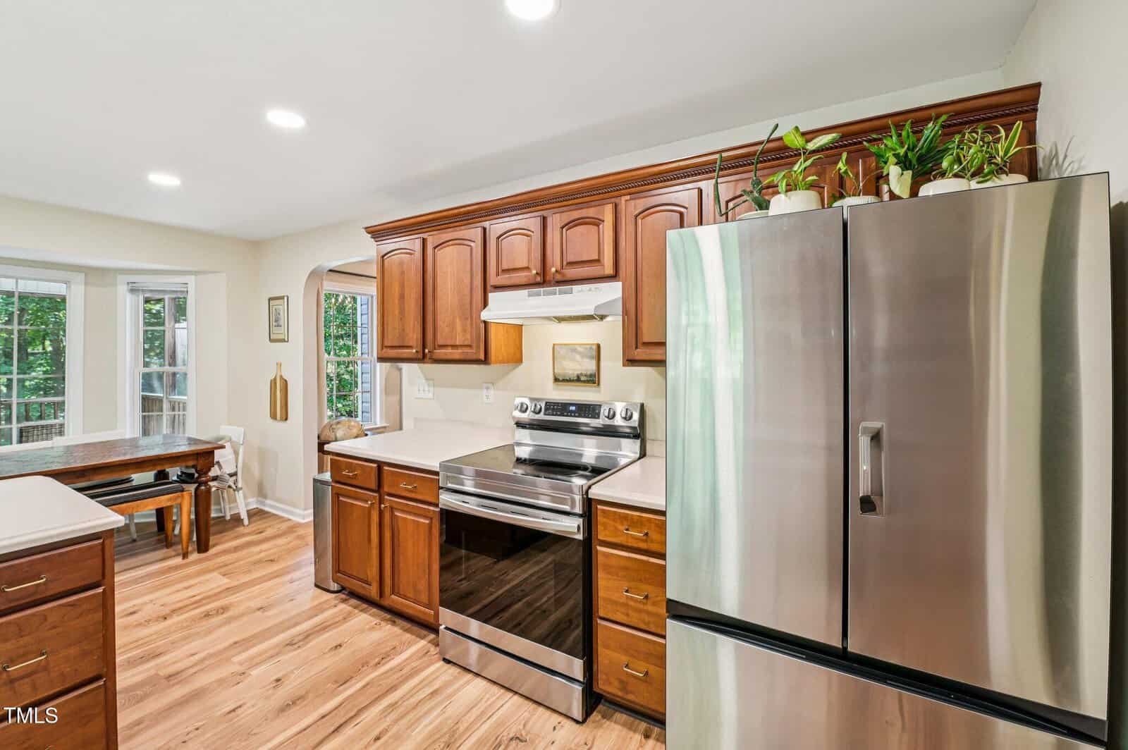 A kitchen with wooden cabinets, stainless steel appliances including a refrigerator and electric stove, light countertops, houseplants on top of the cabinets, wood floors, and a windowed dining nook in the background.