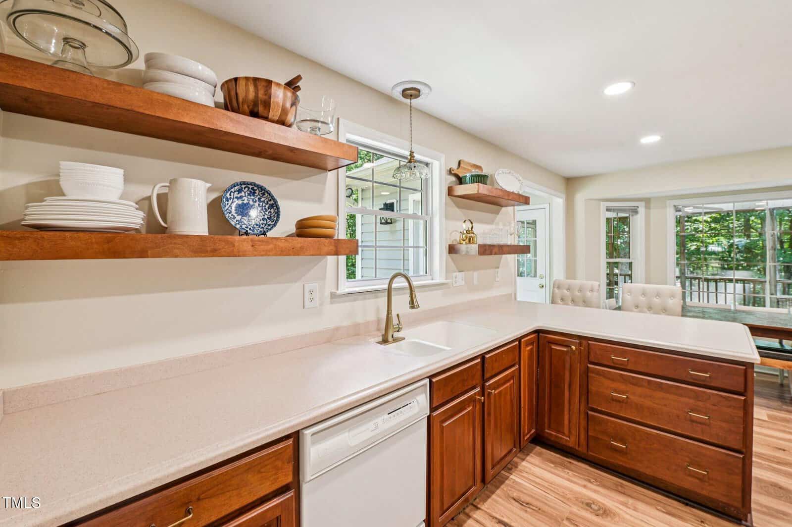 Bright kitchen with wood cabinets, open shelves holding dishes, a white farmhouse sink, brass faucet, and large windows letting in natural light. Dining area with beige chairs and table visible in the background.