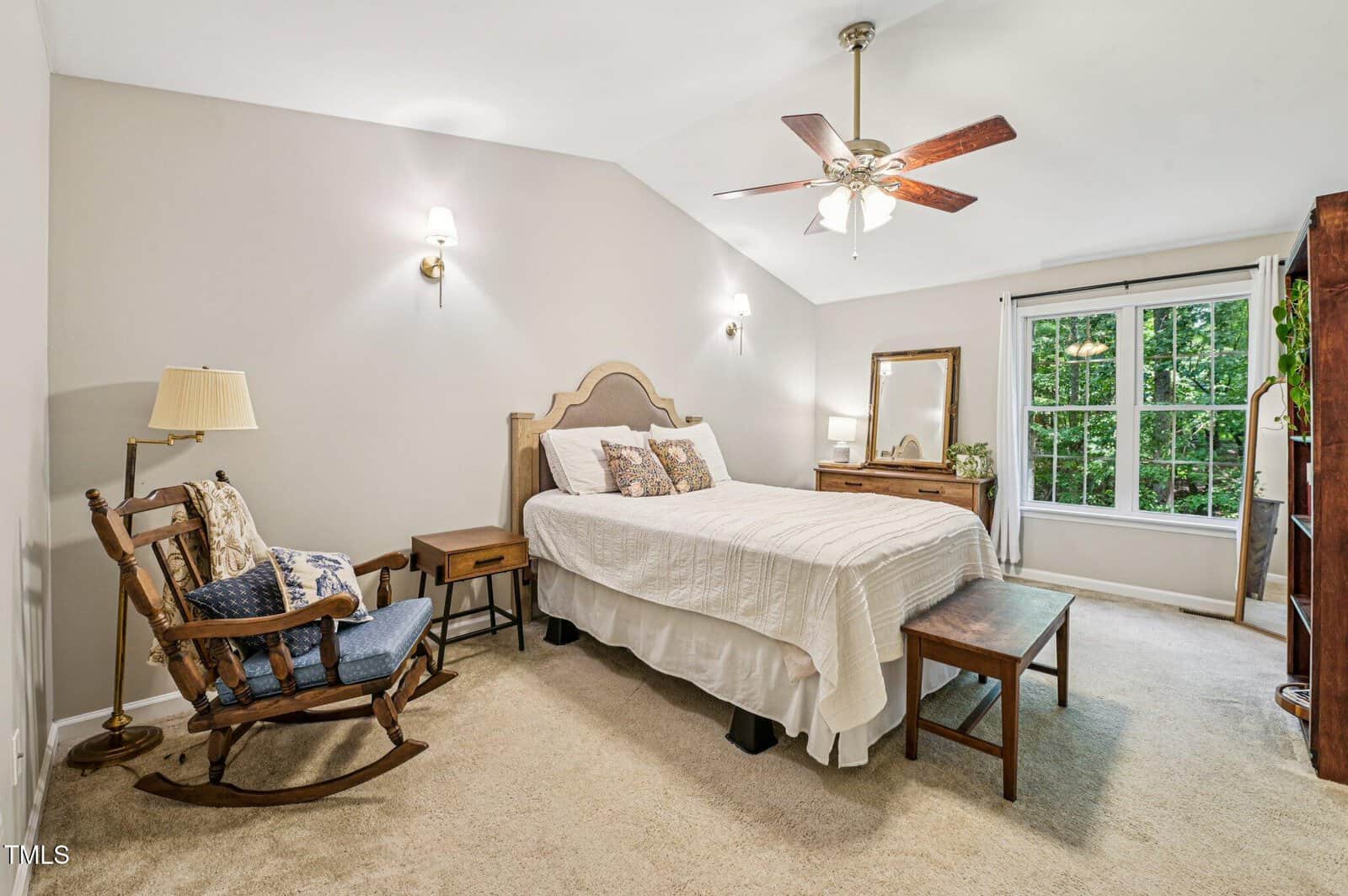 Bright bedroom with a bed, wooden bench, rocking chair, nightstand, dresser with mirror, carpet flooring, ceiling fan, and large window overlooking green trees. Neutral tones and cozy décor create a welcoming atmosphere.