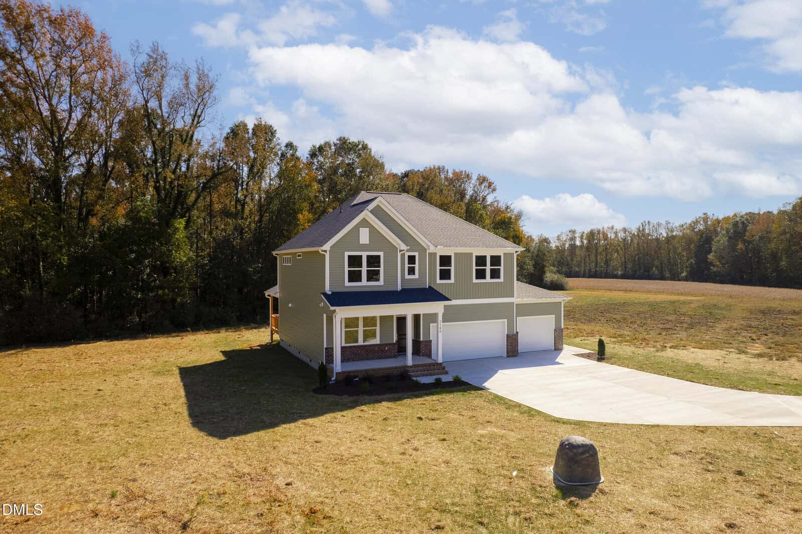 Two-story green house with a front porch and attached three-car garage, situated on a large, open grassy lot bordered by trees under a partly cloudy sky. A curved driveway leads to the garage.