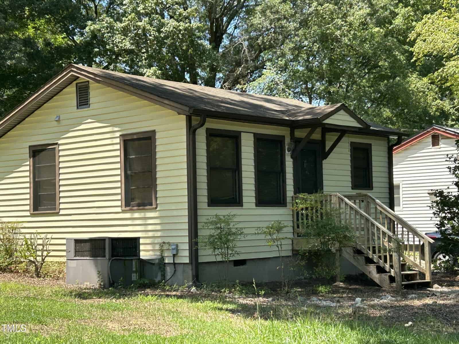A small yellow house with brown trim and a front porch, surrounded by greenery and trees. Wooden steps lead to the front door, and there are several windows on the front and side of the house.