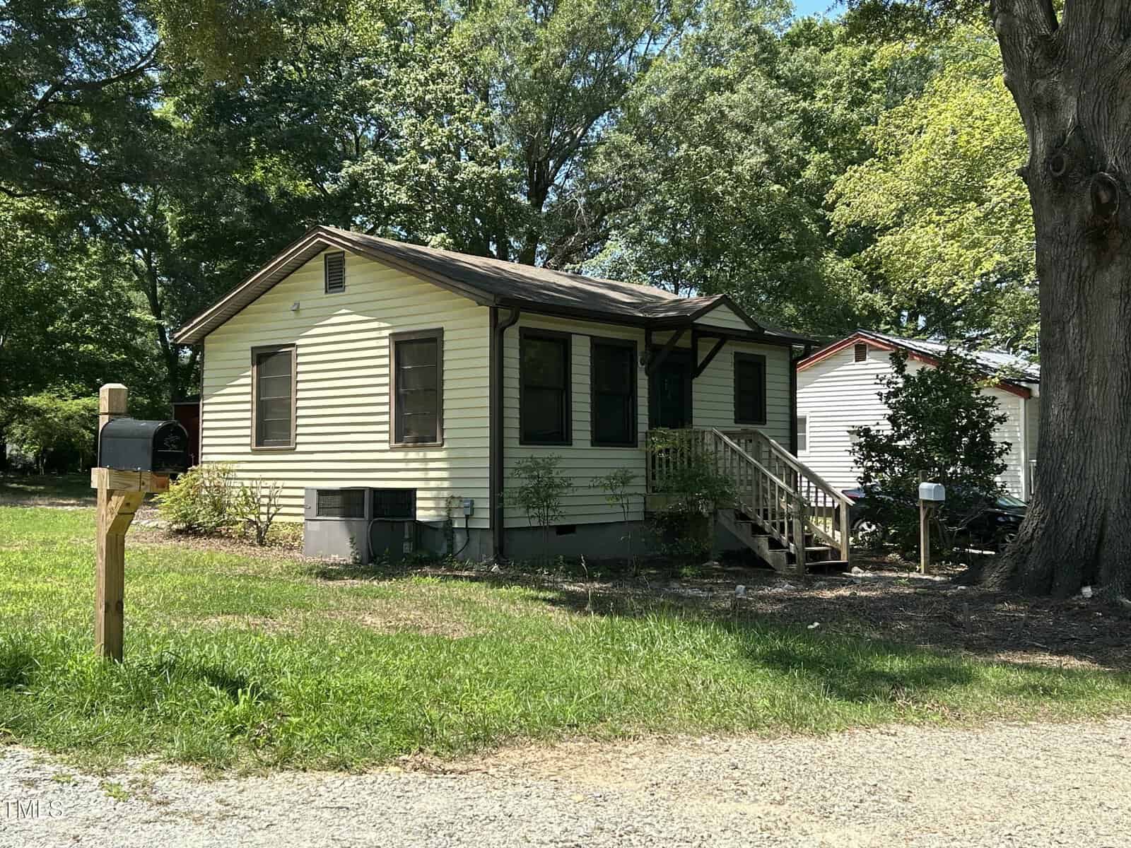 A small, single-story cream-colored house with brown trim and a front porch, surrounded by greenery and trees, sits beside a gravel driveway. A mailbox is on a wooden post near the road.