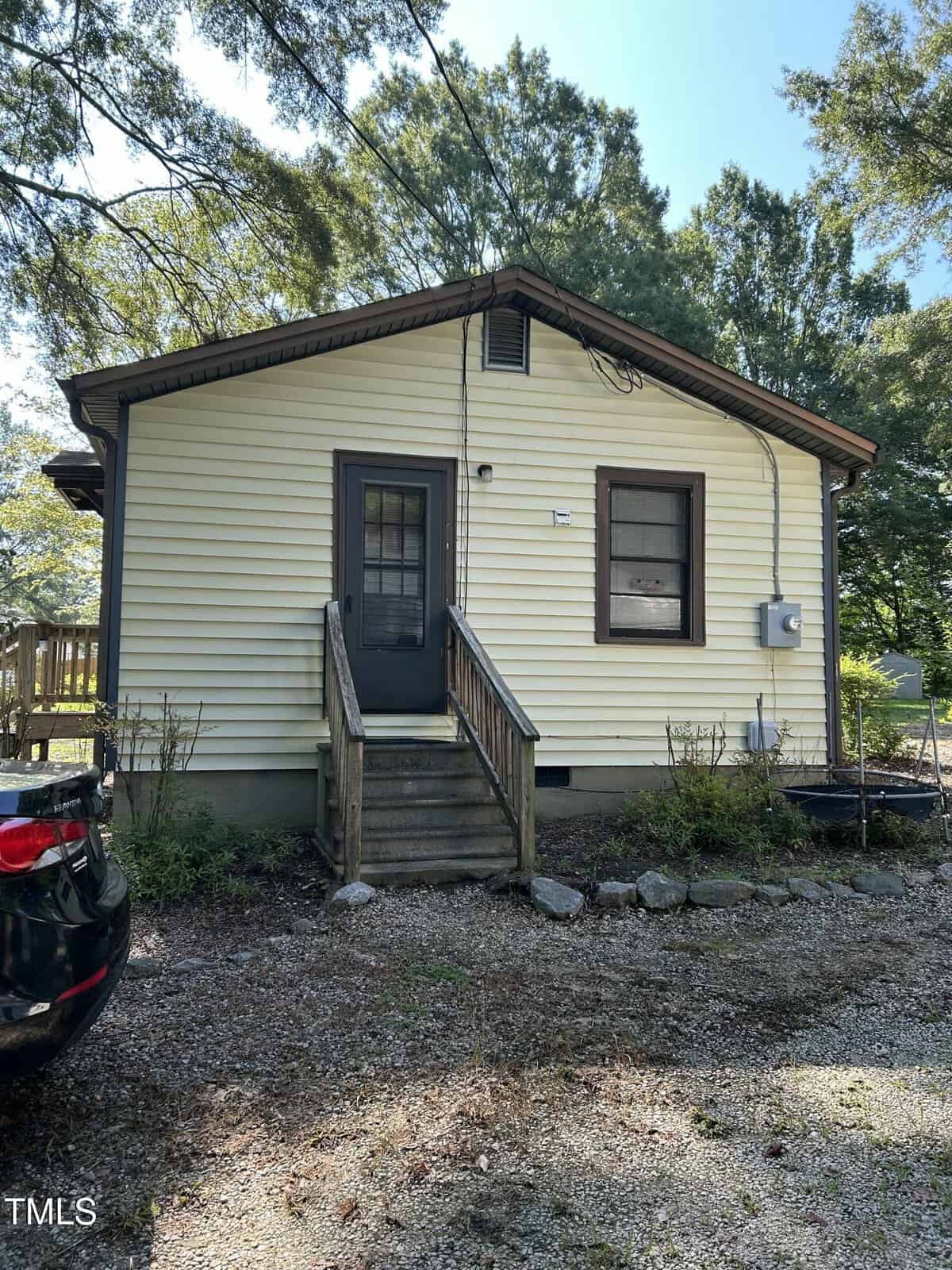 Small yellow house with brown trim, a black door with a window, wooden steps, and a gravel driveway. There are trees in the background and a black car partially visible on the left side of the image.