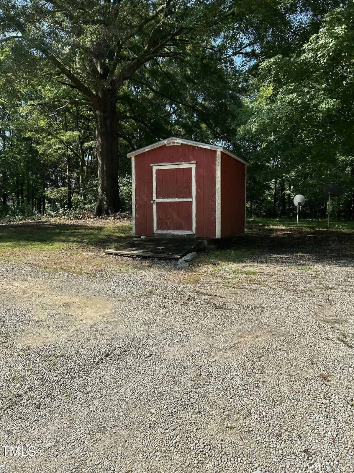 A small red shed with white trim sits on a patch of grass and gravel, surrounded by tall trees and shaded by their foliage on a sunny day.
