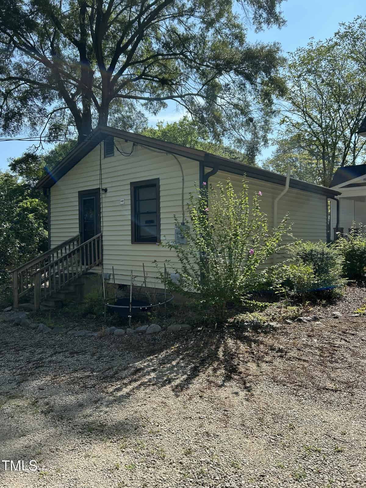 A small yellow house with a sloped roof, wooden steps, and a garden with shrubs and flowers in the front, surrounded by tall trees and a gravel driveway on a sunny day.