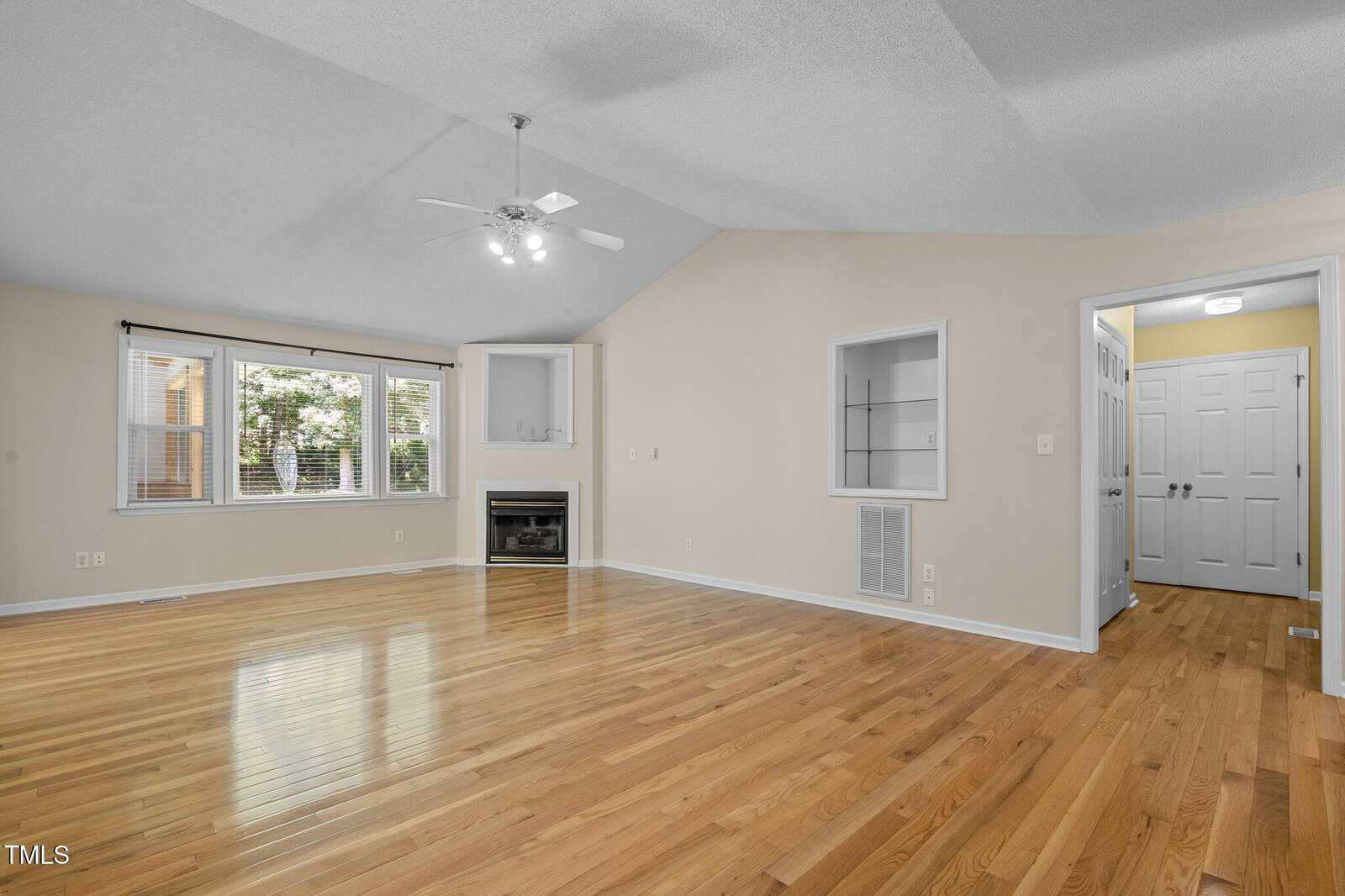 Bright, spacious living room with polished hardwood floors, light beige walls, large windows, a ceiling fan, built-in shelving, and a fireplace in the corner; entryway with double doors visible.