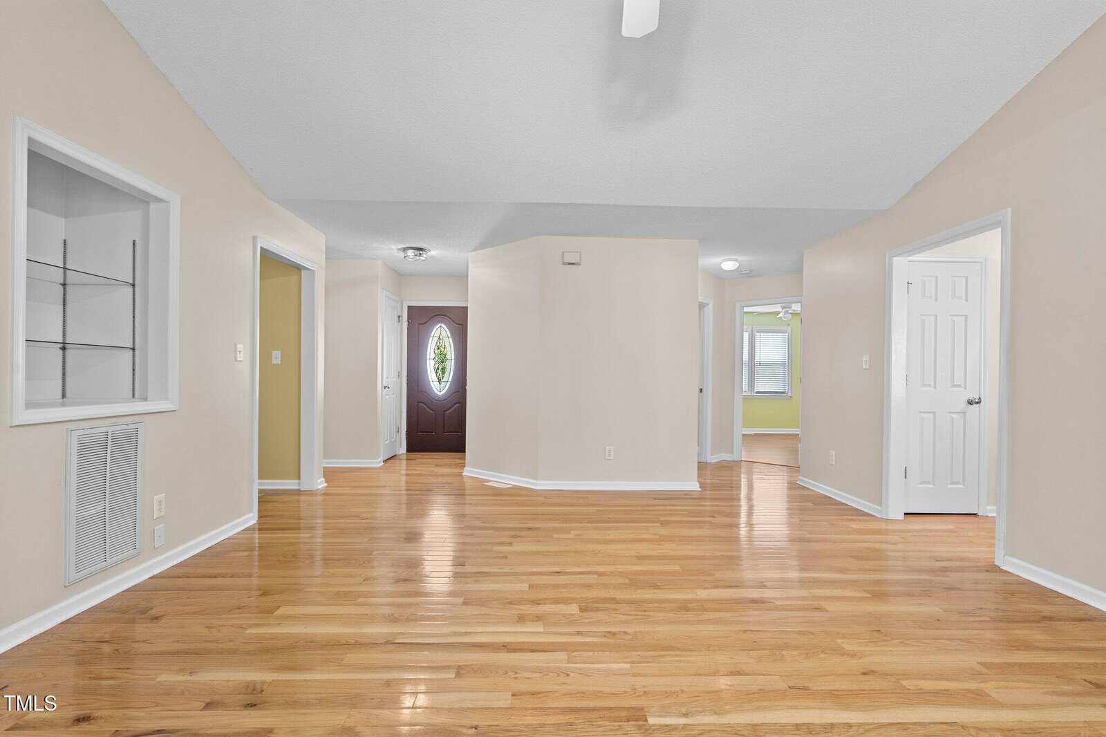 Spacious living area with light hardwood floors, beige walls, white trim, open doorways, built-in shelves, and a dark wood front door with an oval window. Multiple rooms and a hallway are visible.