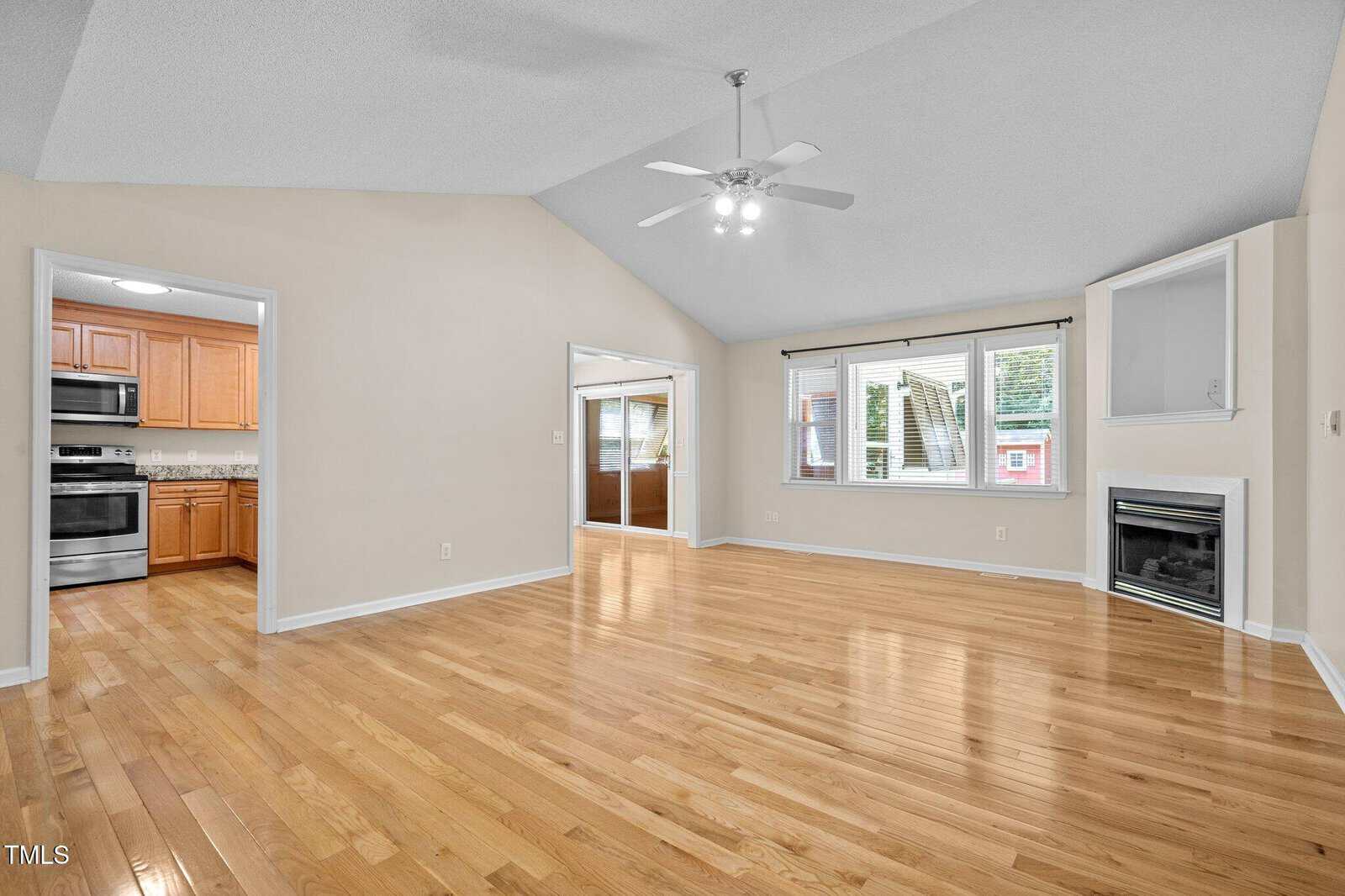 Spacious living room with light hardwood floors, vaulted ceiling, ceiling fan, large window, corner fireplace, and view into a kitchen with stainless steel appliances and wooden cabinets.