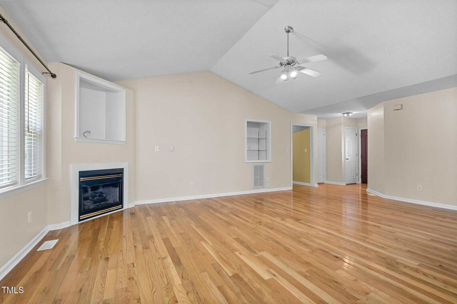 A spacious living room with light hardwood floors, beige walls, large windows with blinds, a white ceiling fan, built-in shelves, and a corner fireplace with a white mantel.