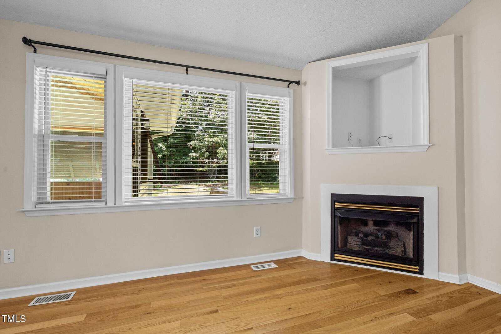 Room with light wood flooring, three large windows with white blinds, a beige wall, a black and white gas fireplace, and a built-in corner nook with two shelves above the fireplace. Trees are visible outside the windows.