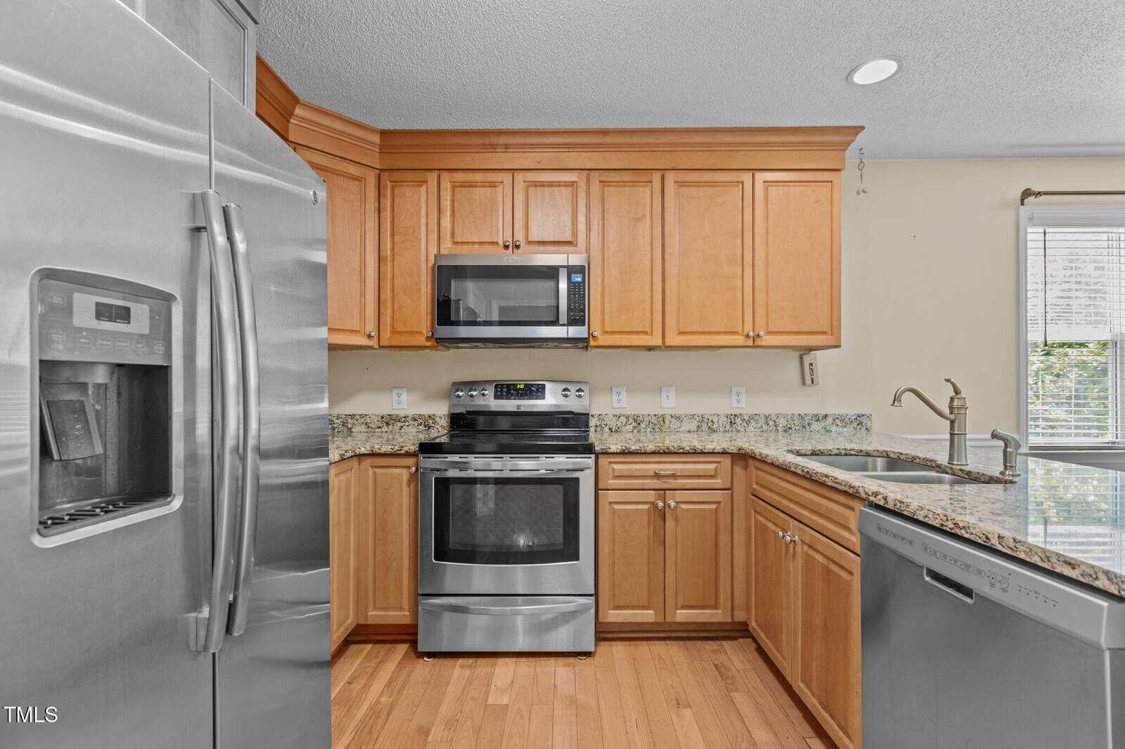 Kitchen with stainless steel appliances, light wood cabinets, granite countertops, a double sink, and a window letting in natural light. The floor is wood, and the kitchen includes a refrigerator, oven, microwave, and dishwasher.