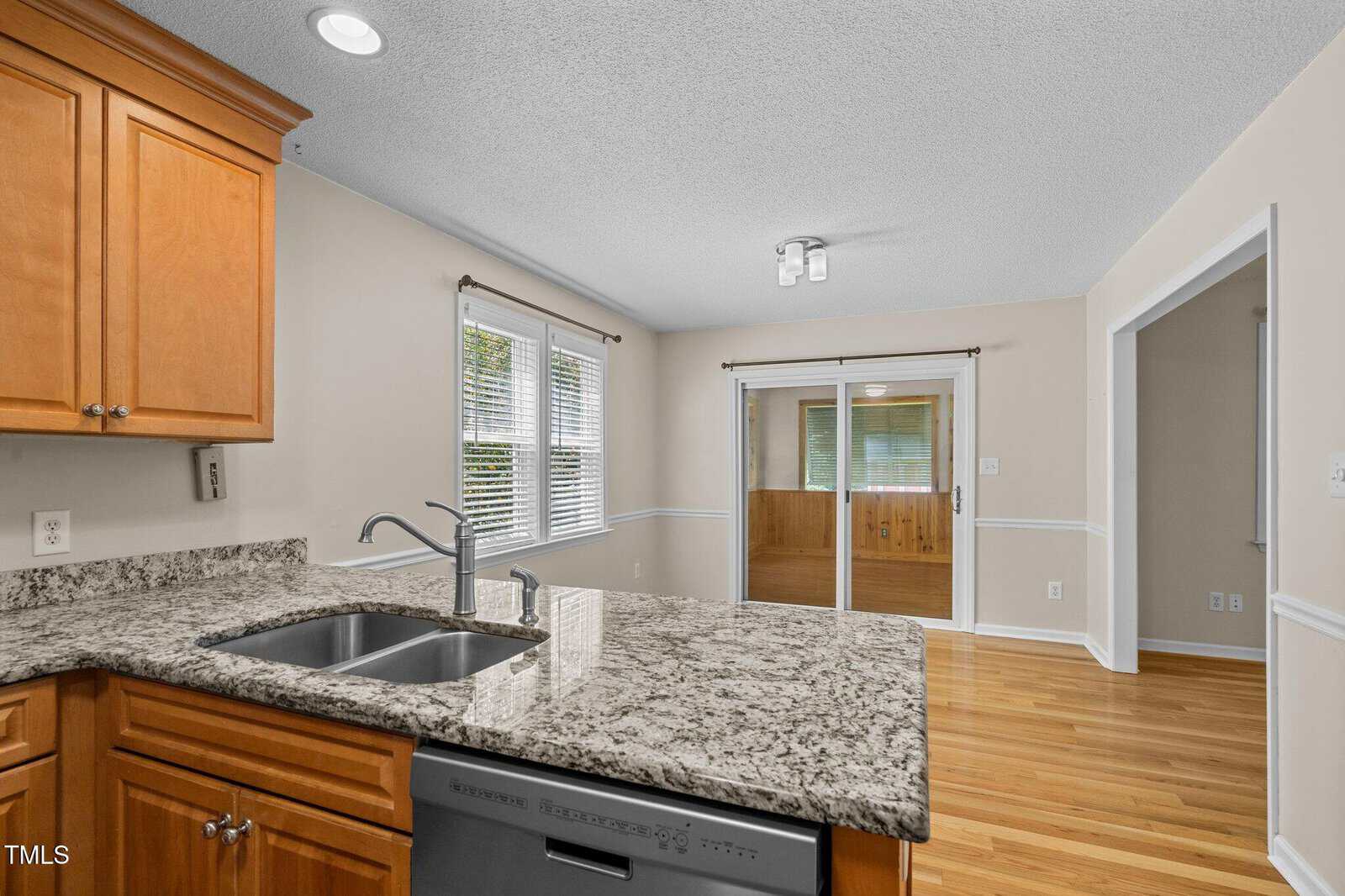 Kitchen with granite countertops and wood cabinets overlooks a dining area with hardwood floors, neutral walls, two windows, and sliding glass doors leading to a sunroom. Ceiling light fixtures visible in both rooms.