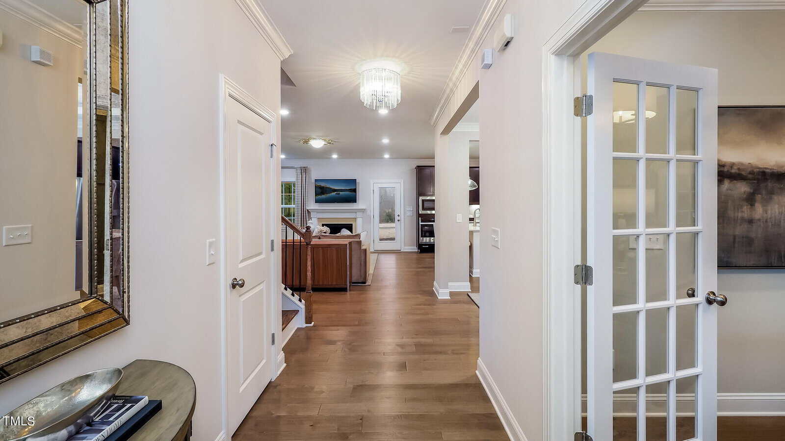 Bright hallway with light wood floors, neutral walls, and a glass-paned door. The hallway opens to a living area with modern furniture, artwork, a chandelier, and a large mirror on the left wall.