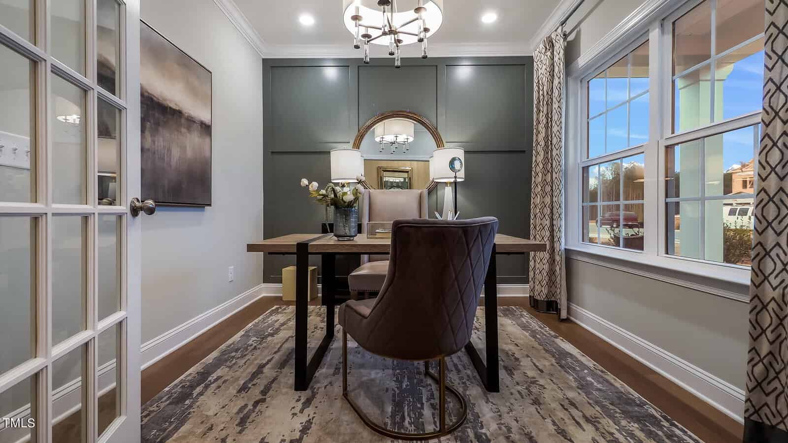 A modern home office with a large wooden desk, two chairs, a table lamp, decorative mirror, and vases. The room has green accent walls, patterned curtains, a rug, and large windows letting in natural light.