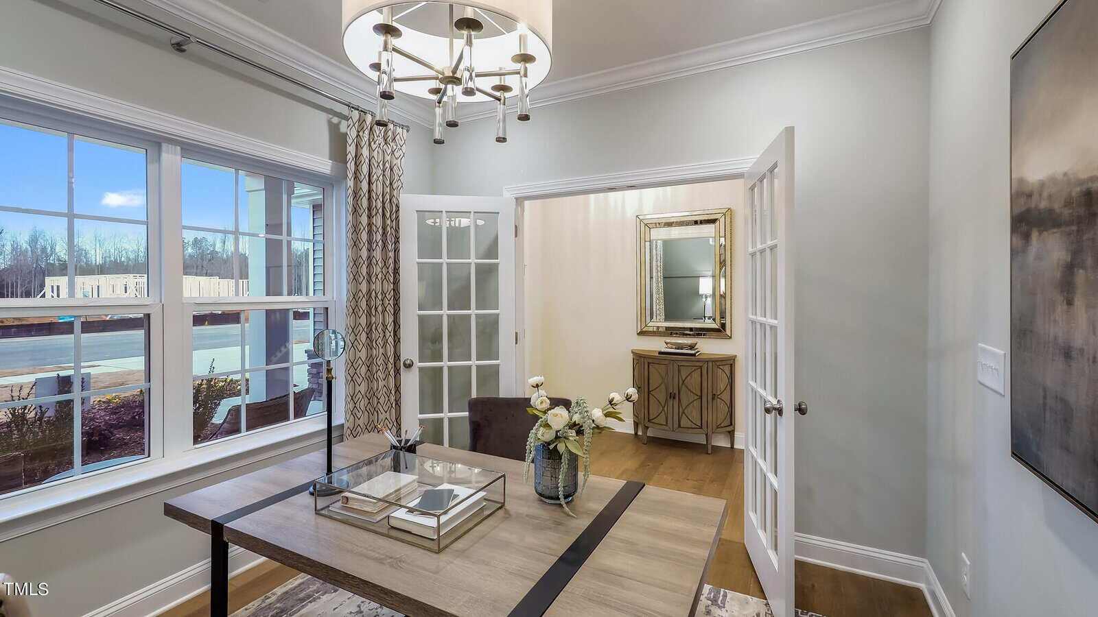 A modern home office with a wooden desk, a brown chair, and a vase of white flowers, set near large windows overlooking a backyard with a pool. French doors open to a hallway with a mirror and console table.