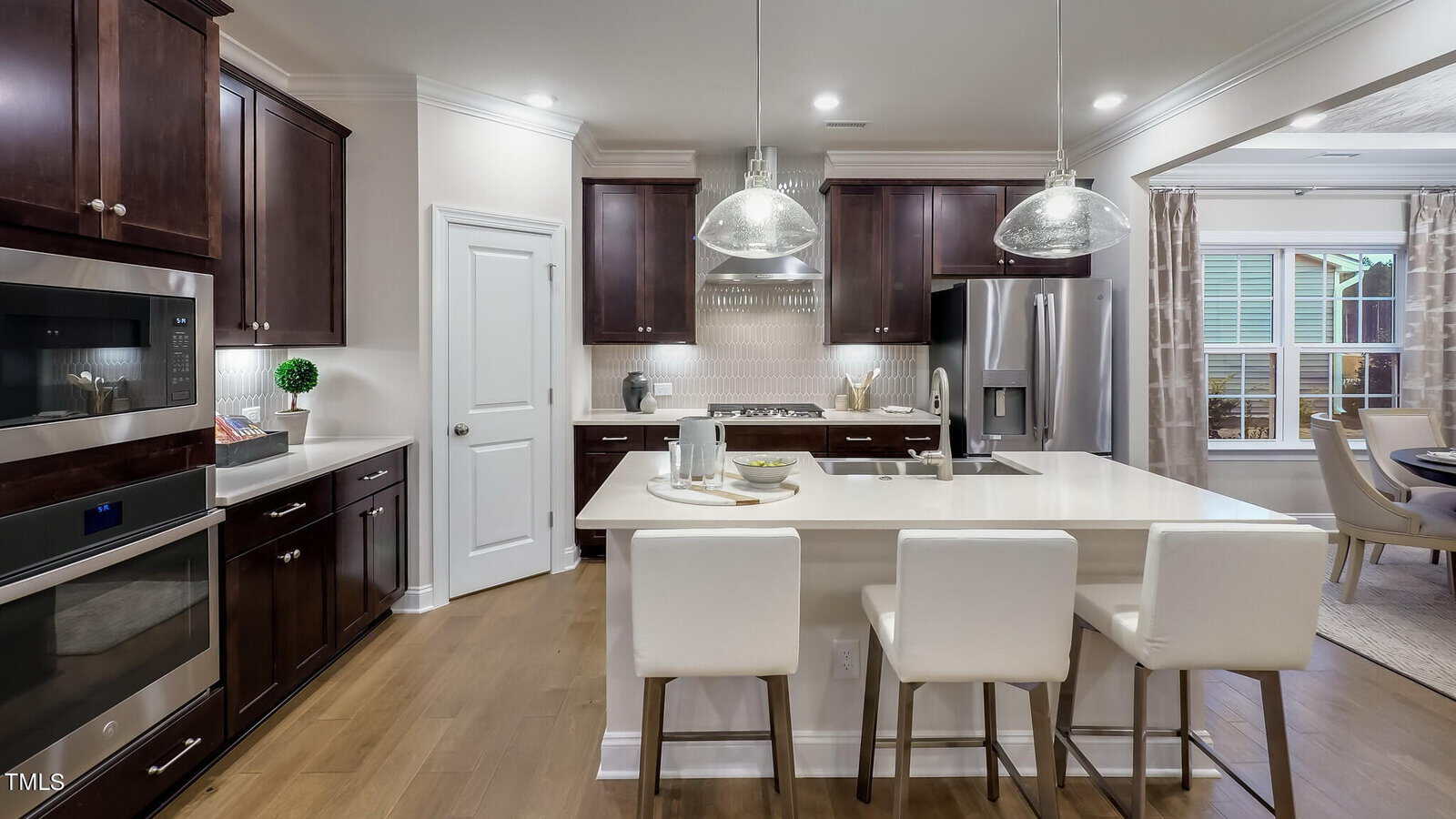 Modern kitchen with dark wood cabinets, stainless steel appliances, white island with three white chairs, pendant lights above, and a view into a dining area with a table and chairs through an open doorway.