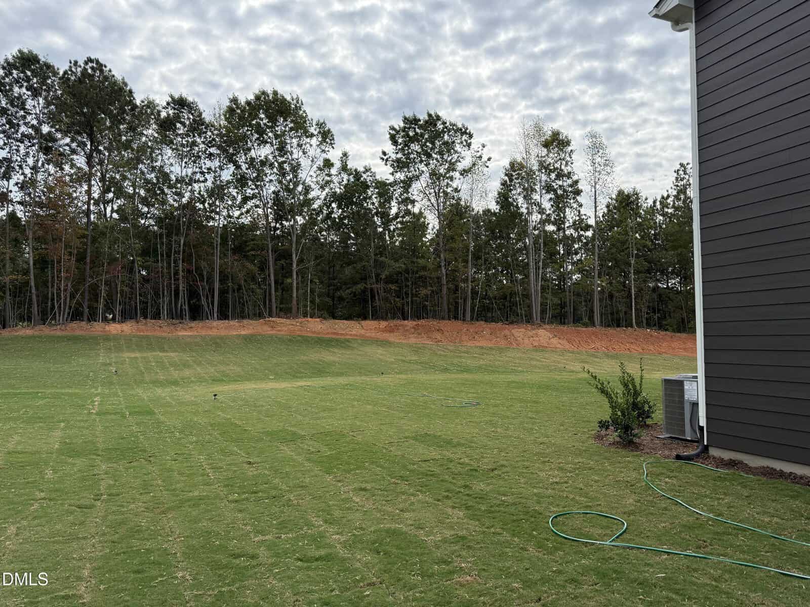 A newly sodded backyard with a green lawn, a hose on the ground, an air conditioning unit by the house, and a tree line at the edge of the property under a partly cloudy sky.