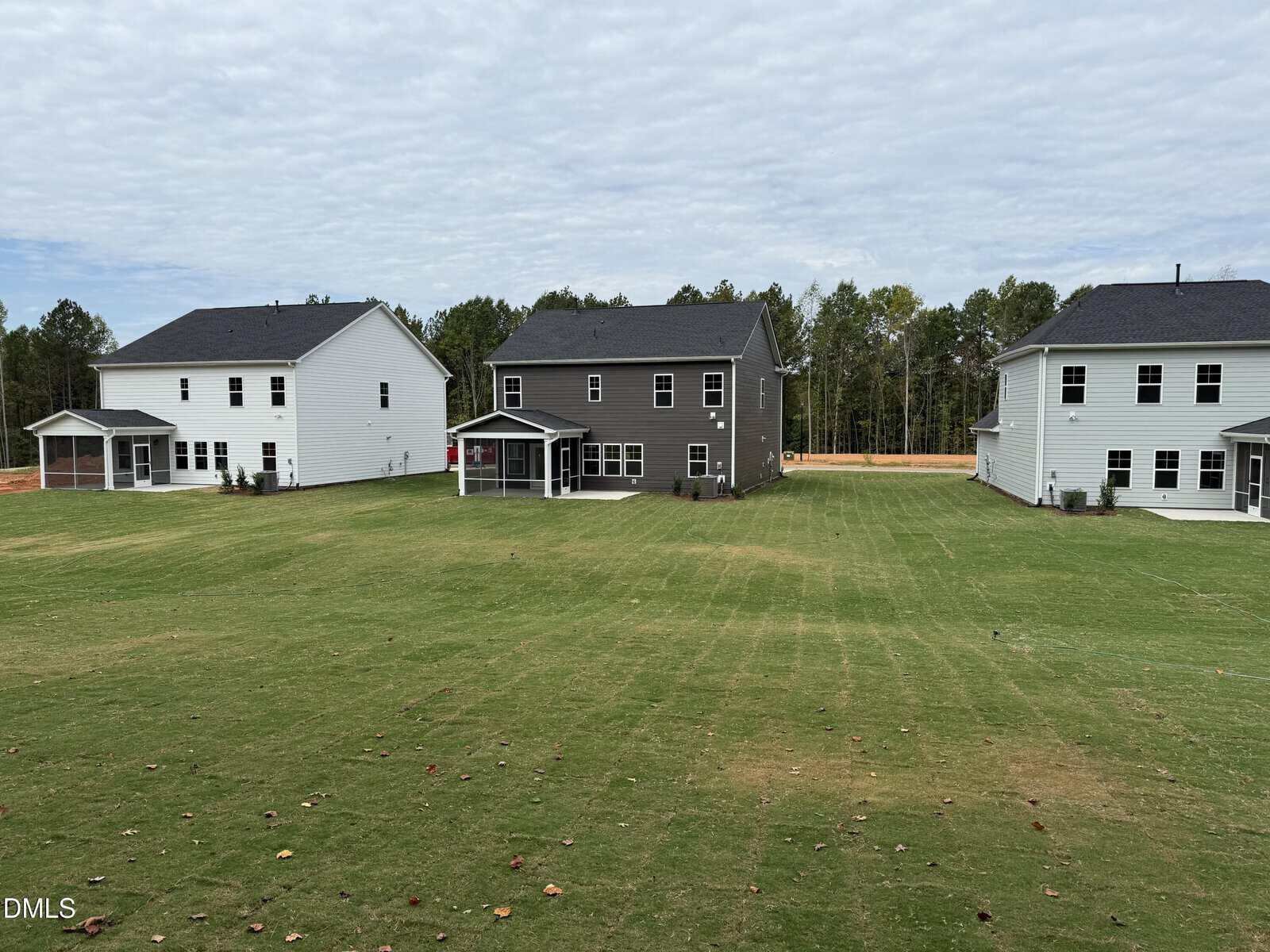 Three two-story houses with covered patios sit on a large, grassy lawn under a cloudy sky. Trees line the background. The houses are white and dark gray, all with multiple windows.