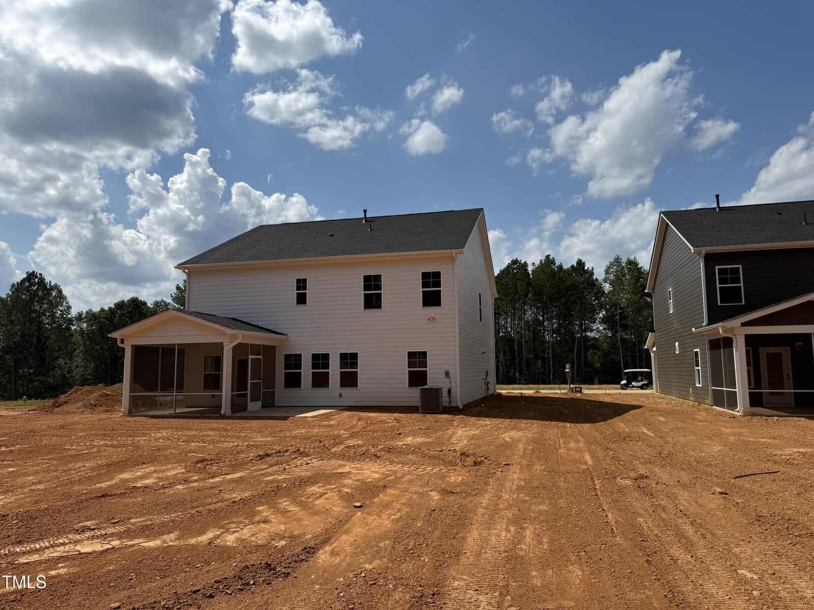 Two new houses with white and dark siding stand on red dirt lots under a partly cloudy sky; the yard is unfinished with no grass or landscaping, and trees are visible in the background.