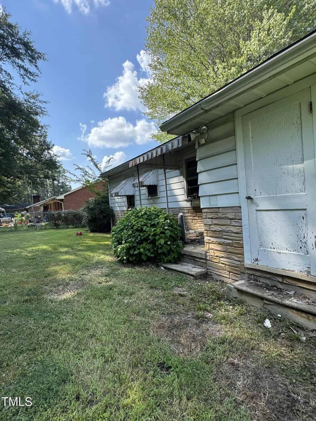 A weathered house with white siding and stone accents, featuring a bush near the entrance, sits on a grassy yard under a partly cloudy sky. The door and exterior appear worn and in need of maintenance.