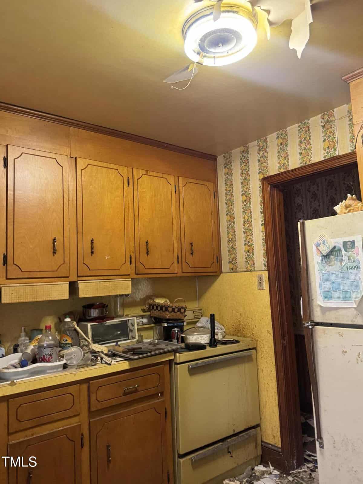 Cluttered vintage kitchen with wooden cabinets, yellow stove, worn wallpaper, white refrigerator, and debris on the floor. A broken ceiling fan with exposed blades hangs above.