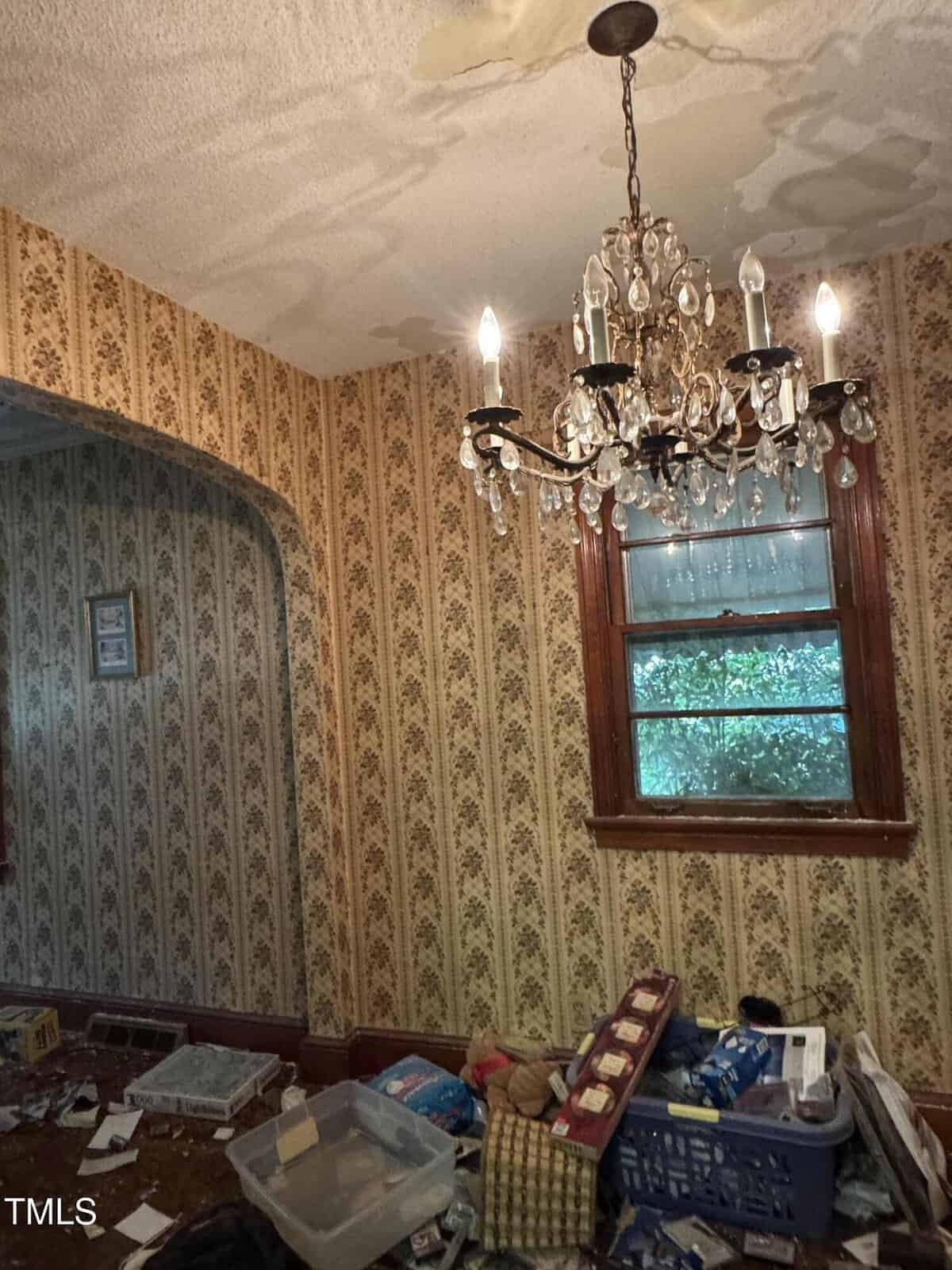 A cluttered room with vintage wallpaper, a crystal chandelier, a window with a wooden frame, and various boxes and items scattered on the floor. The ceiling shows signs of water damage.