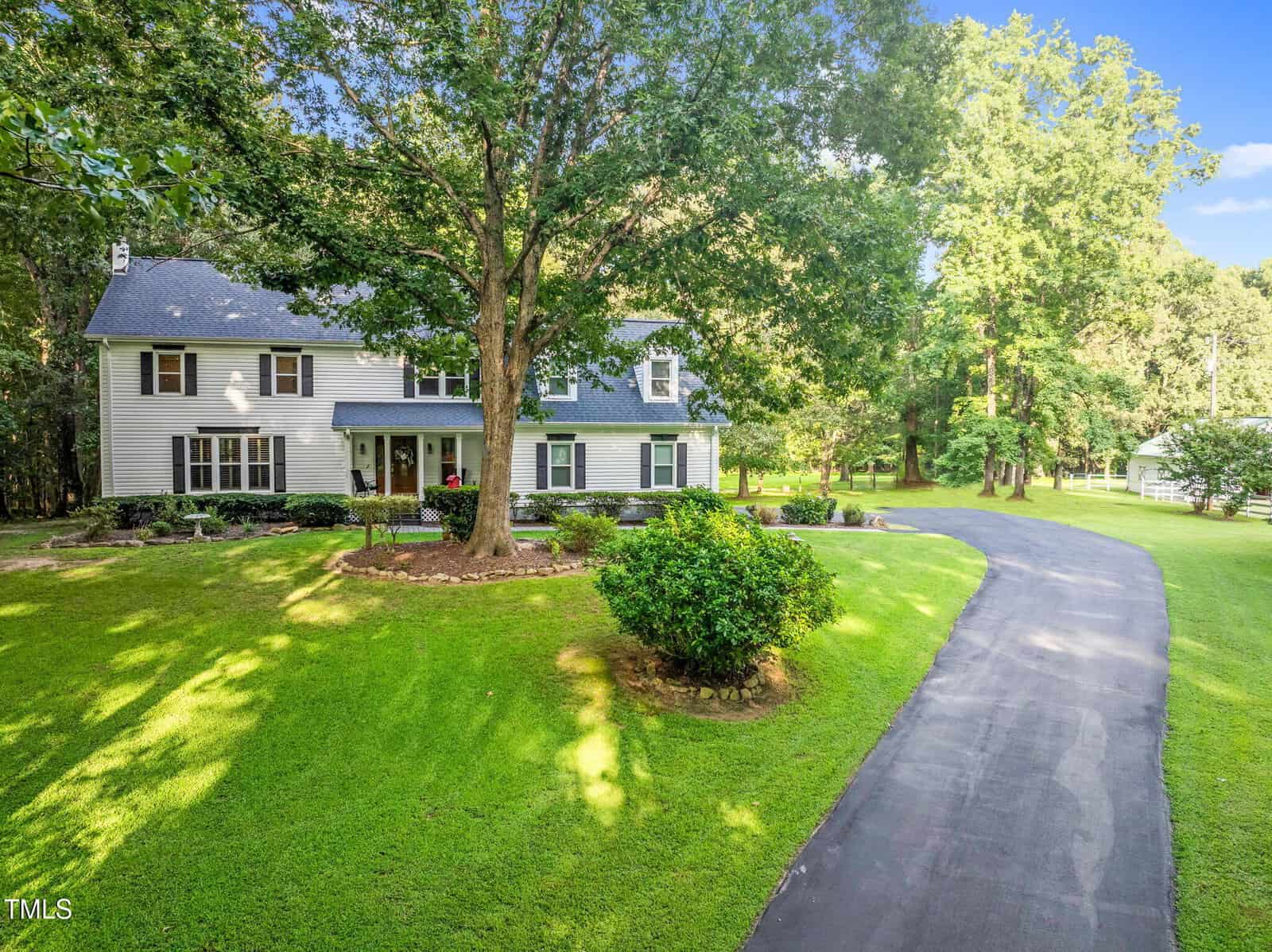 A two-story house with gray siding and dark shutters, set among green grass and trees, awaits you at 687 Cooke Road in Louisburg. A curved paved driveway leads to the home, with landscaped bushes enhancing the front yard’s charm.