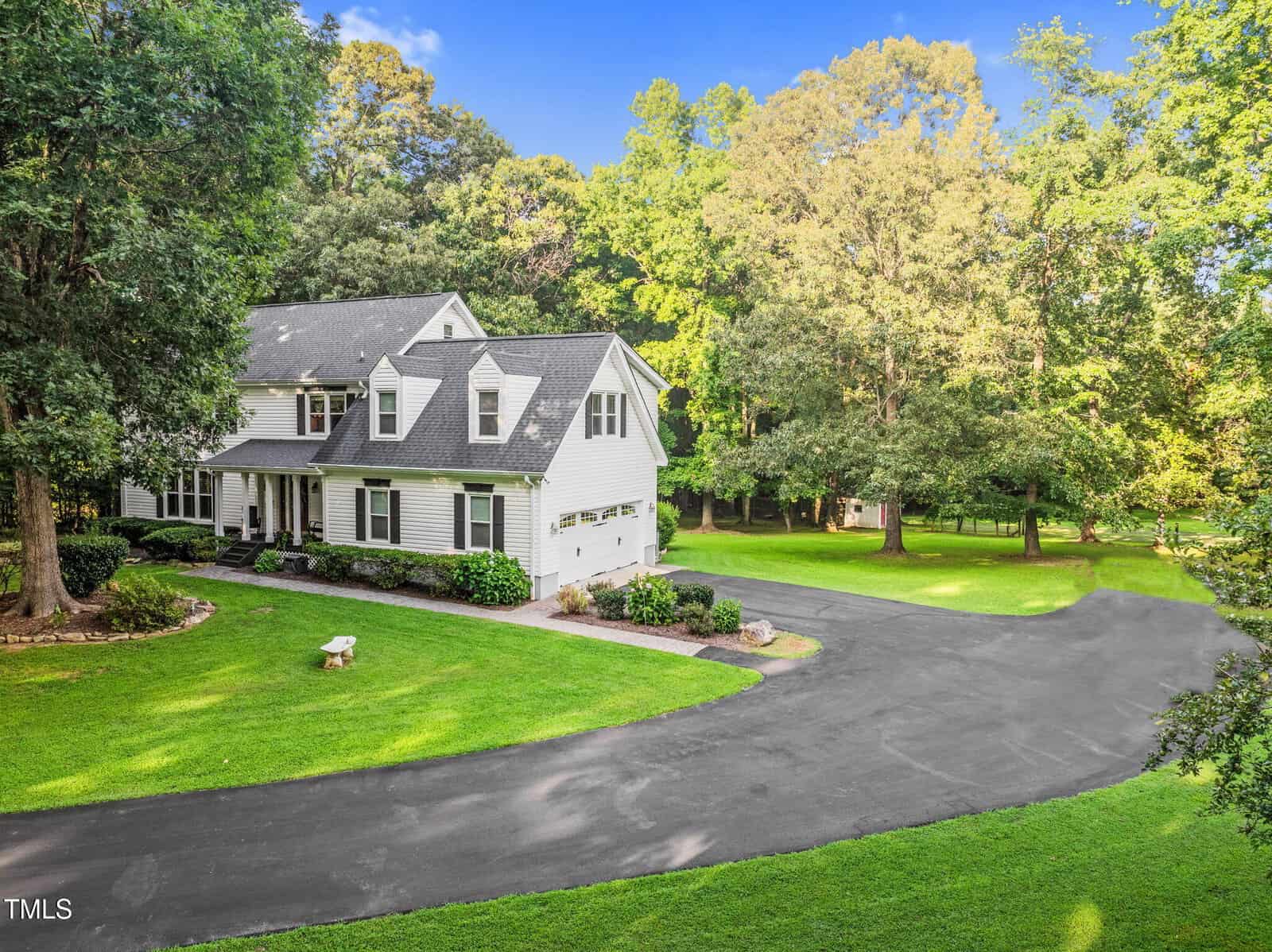 Two-story white house with gray roof, attached three-car garage, and large paved driveway, surrounded by green lawn, mature trees, and bright landscaping on a sunny day.