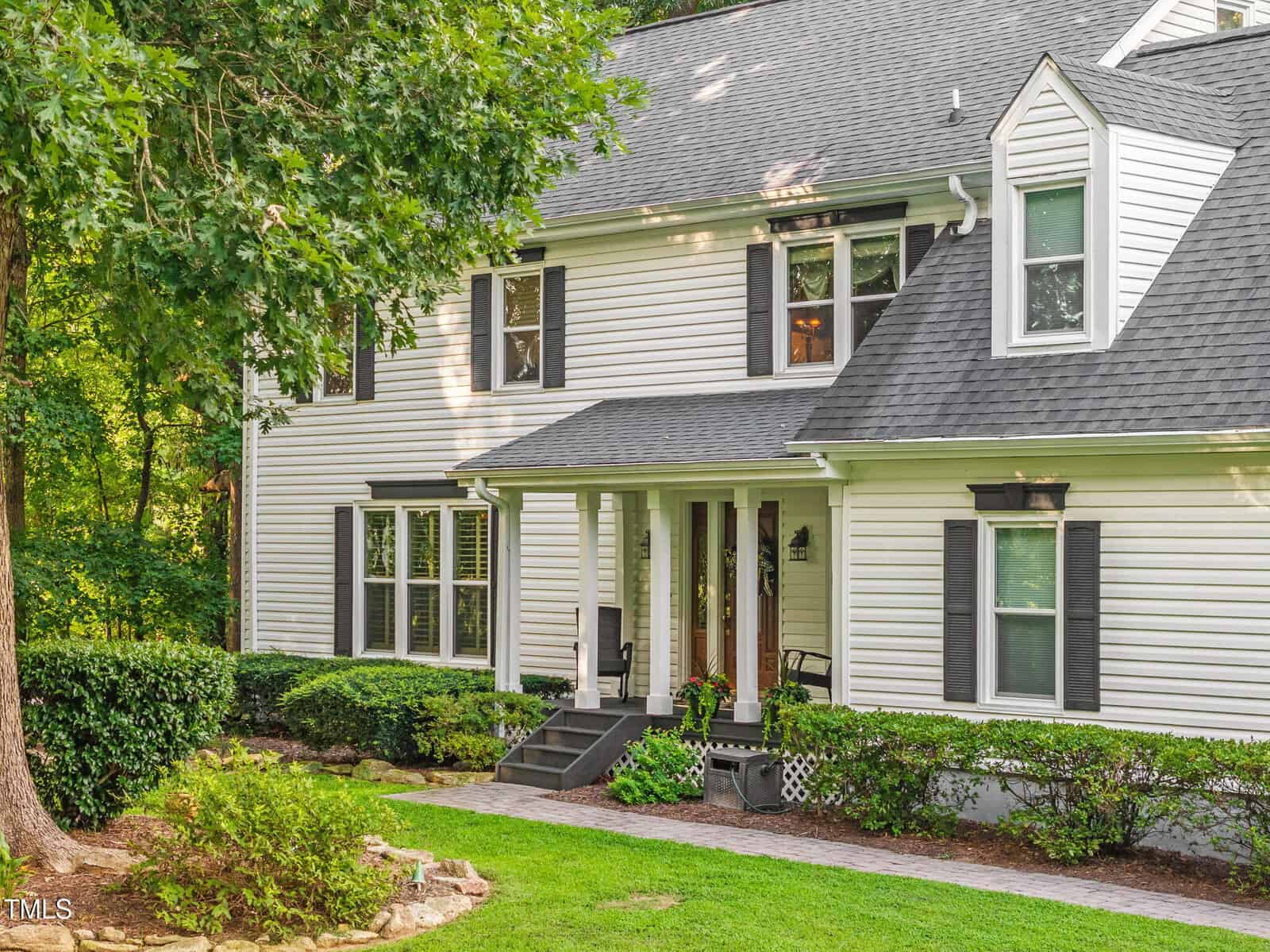 Two-story white house with black shutters, a gray shingle roof, covered front porch, and a brick walkway. The yard has green grass, shrubs, and mature trees in the background. It is sunny with bright lighting.