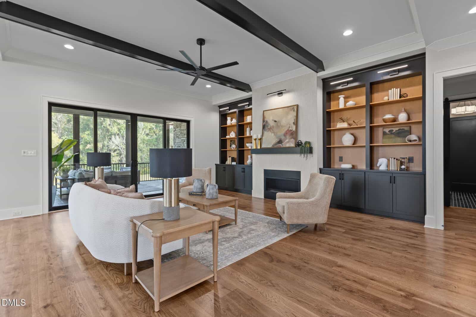 Modern living room with wood floors, white sofa, beige chairs, a coffee table, built-in black shelves with decor, a fireplace, large windows, and black ceiling beams.