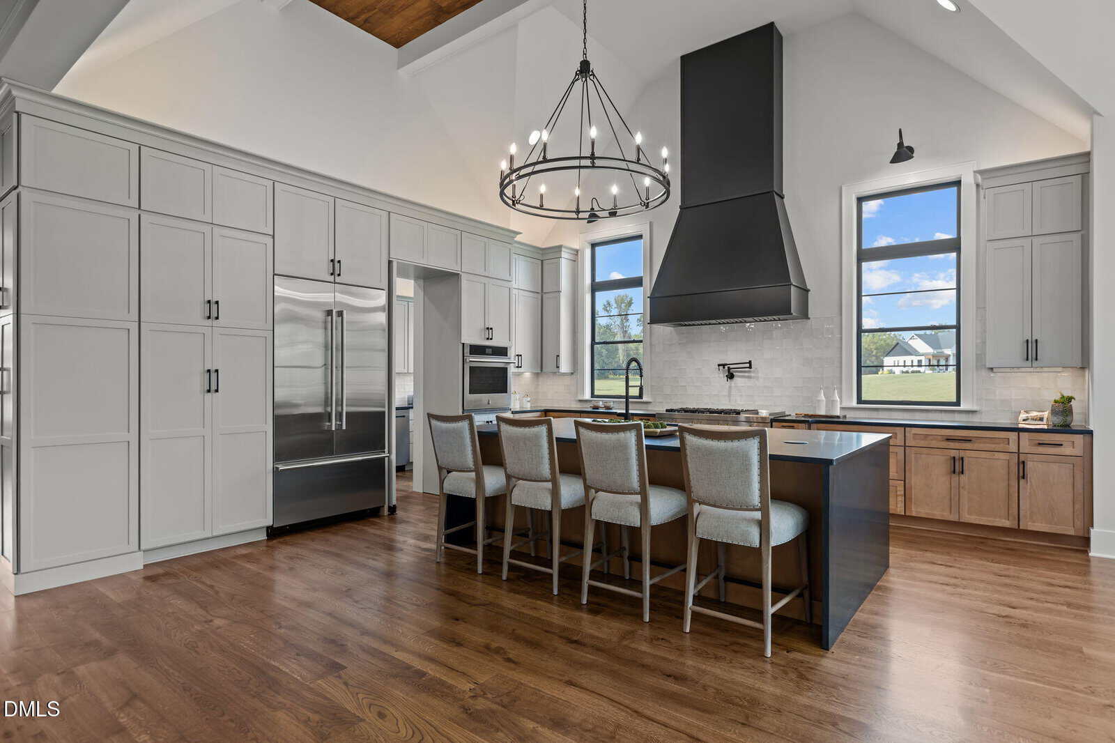 Modern kitchen with light gray cabinets, a large black island with four upholstered barstools, stainless steel appliances, wood flooring, a black range hood, and large windows showing a scenic outdoor view.