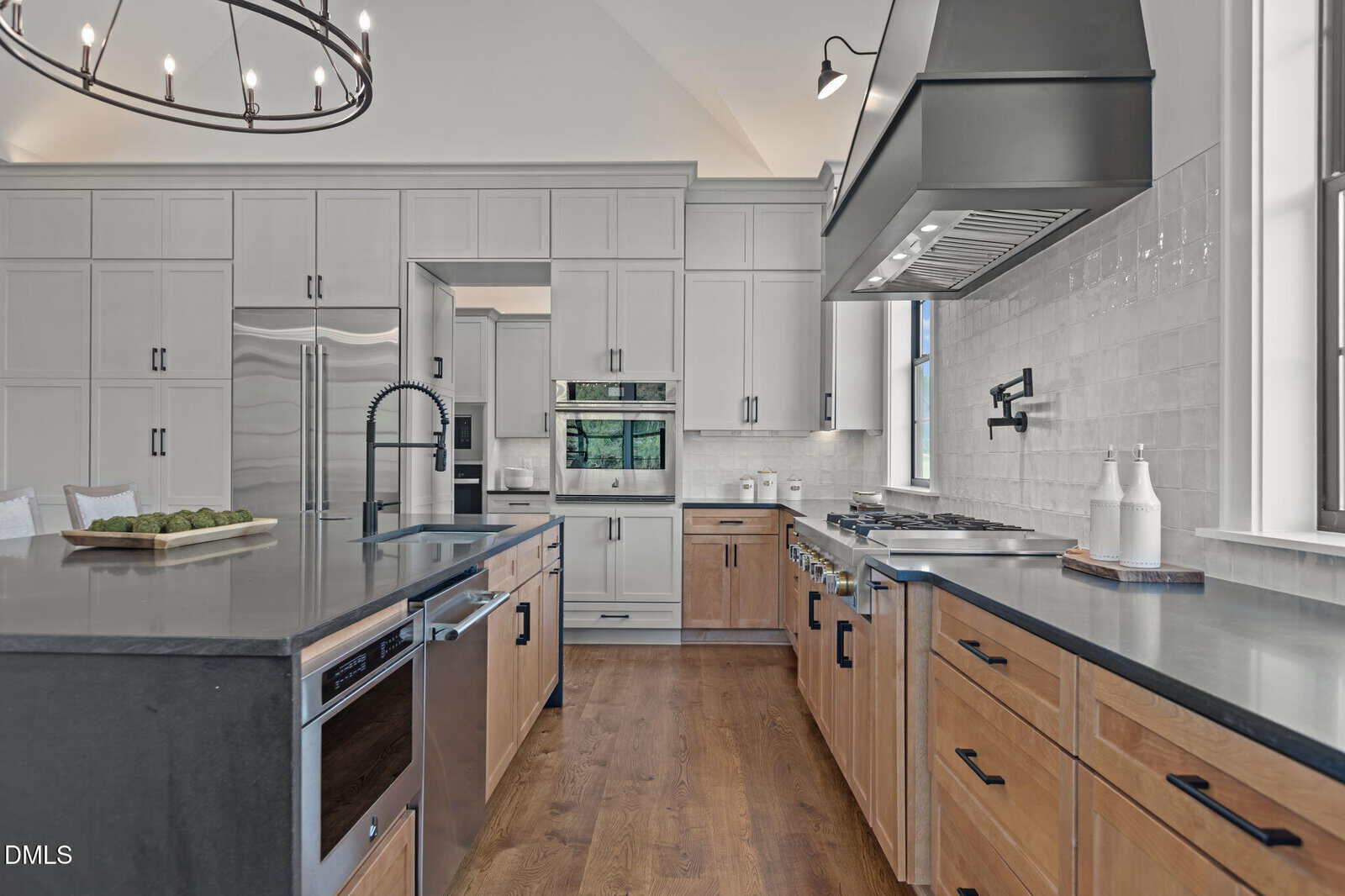 Modern kitchen with light gray upper cabinets, wooden lower cabinets, stainless steel appliances, a large dark island with a built-in sink, and a wood floor. A black chandelier hangs above the island.