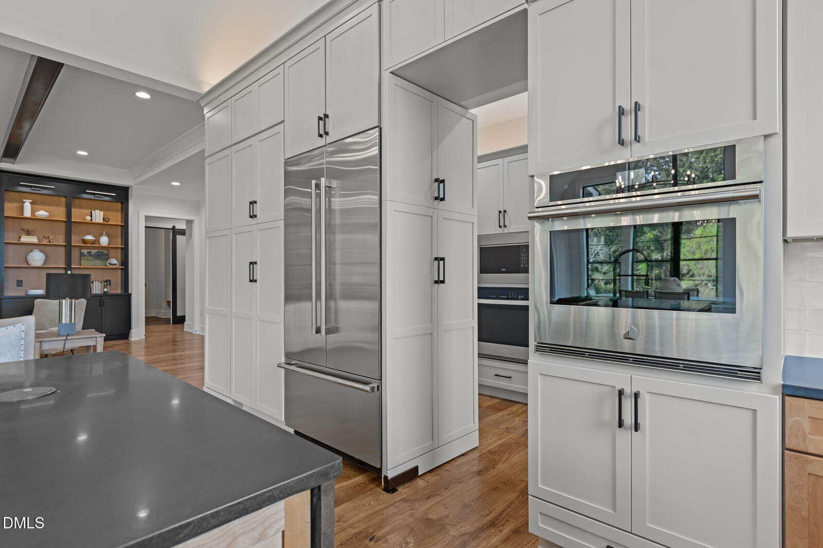 Modern kitchen with white cabinets, stainless steel refrigerator, built-in double oven, dark countertops, and light wood floors. Open shelves and large windows visible in the background.