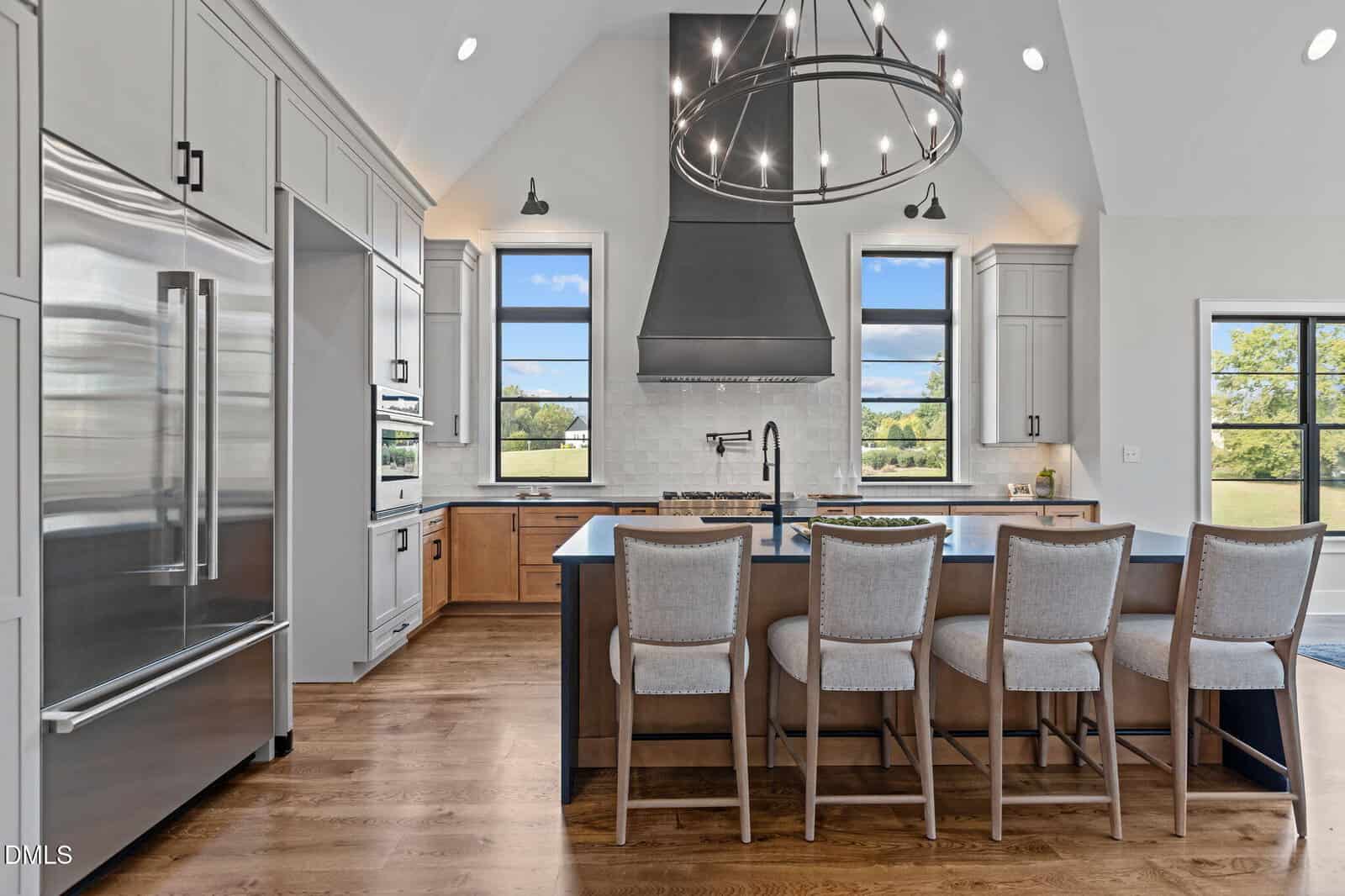 Modern kitchen with a large island, four beige chairs, stainless steel appliances, black range hood, pendant light, wood flooring, white cabinets, and windows showing a green landscape.