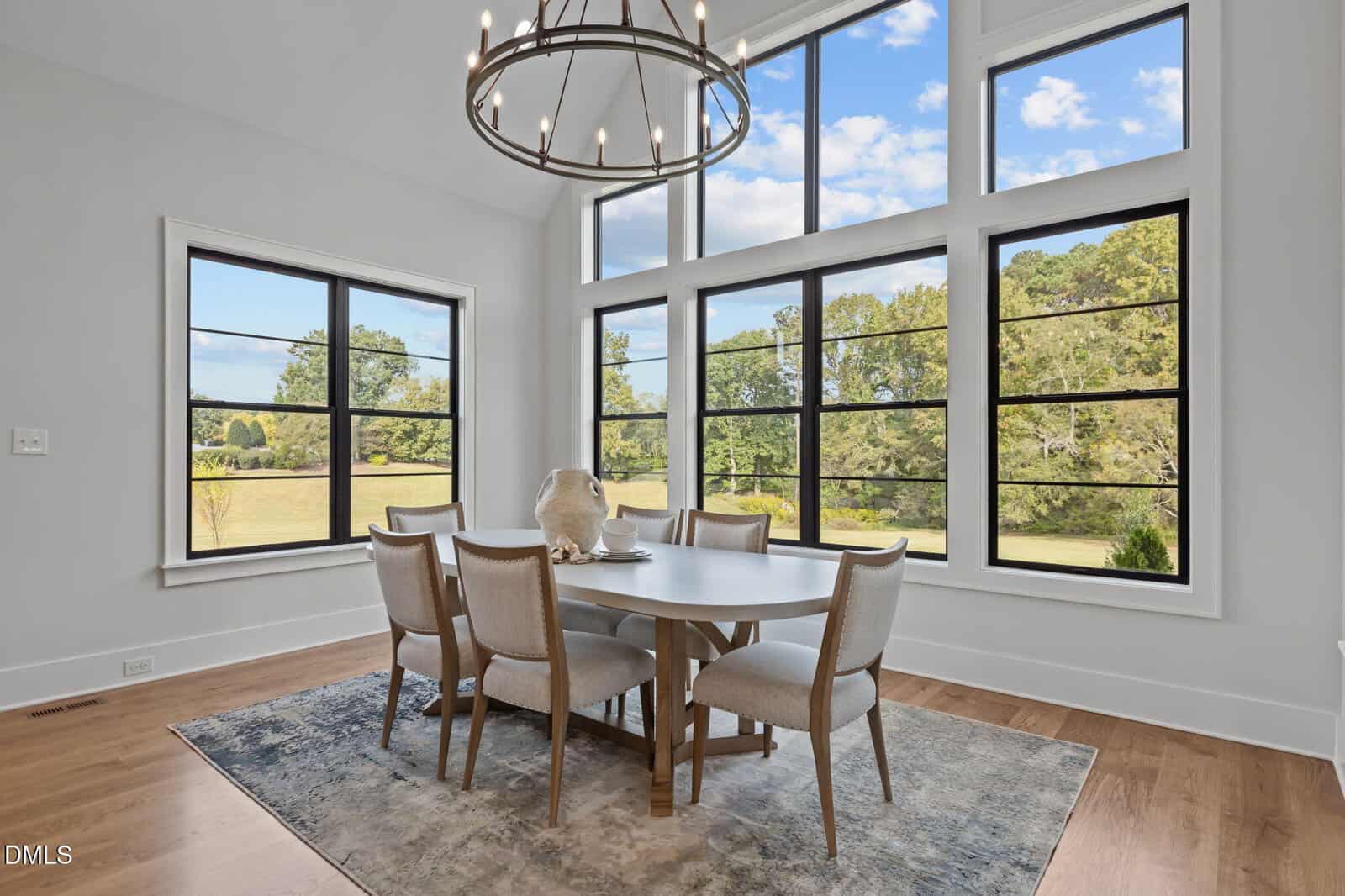 Dining area with six chairs around a round table, large windows with a scenic outdoor view, modern chandelier, light walls, and wooden floor with a patterned rug.