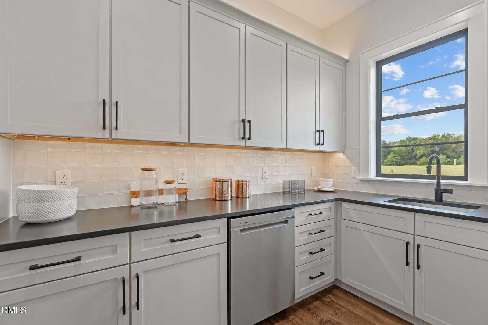 Modern kitchen with white cabinets, black countertops, a stainless steel dishwasher, and a large window with a scenic outdoor view. Decorative jars and a speaker sit on the counter. Light wood flooring throughout.