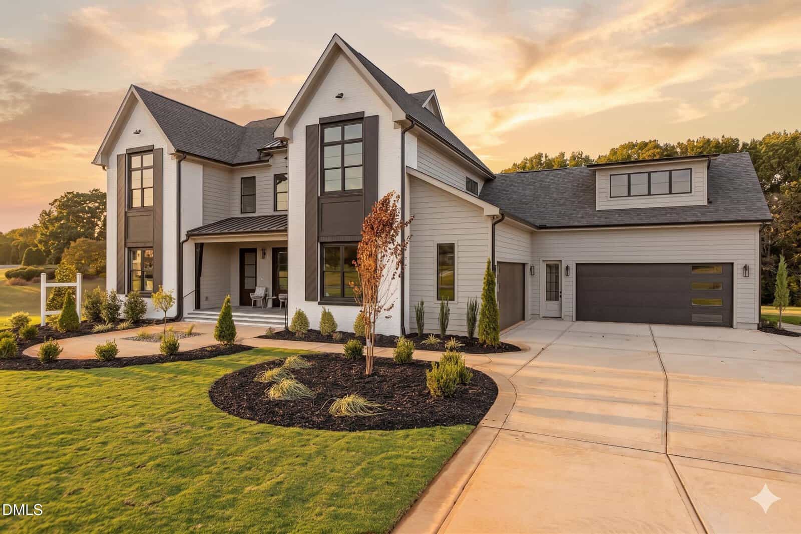 Modern two-story house with large windows, gray and white exterior, attached three-car garage, landscaped front yard, and a clean driveway, set against a sunset sky and surrounded by green grass and trees.