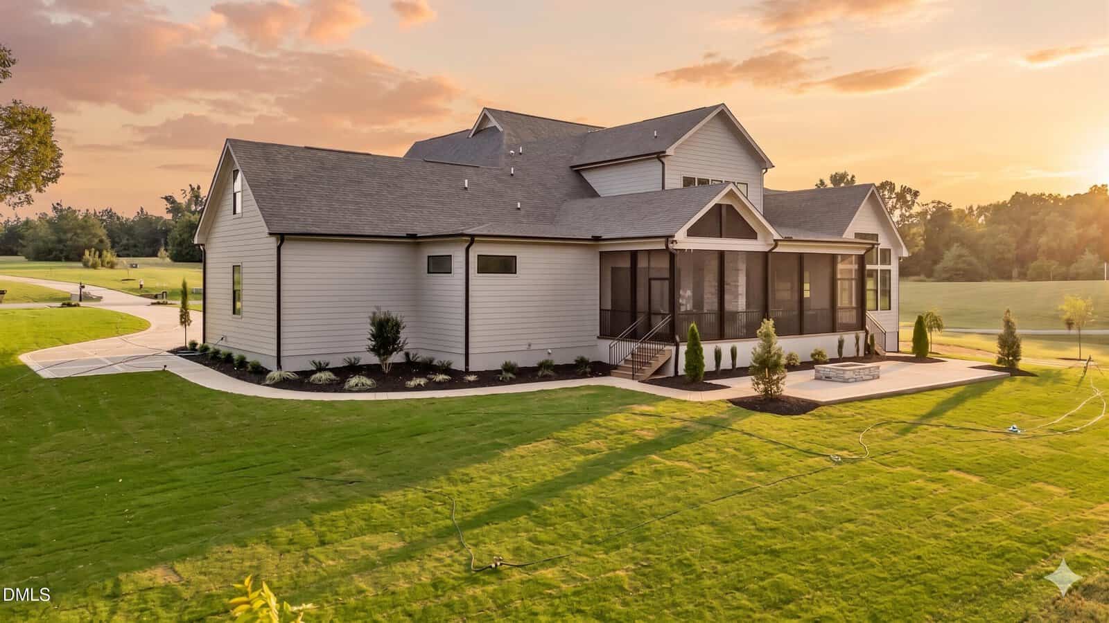 A modern two-story house with gray siding, large windows, and a screened-in porch, set on a spacious, manicured lawn at sunset with trees and a curved driveway in the background.
