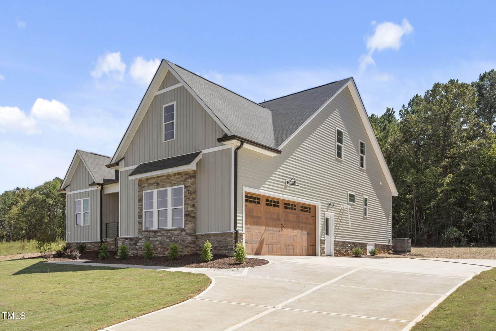 A modern, two-story house with light gray siding, stone accents, and a wooden double garage door. The home is surrounded by a neatly landscaped lawn, a wide driveway, and trees in the background under a blue sky.