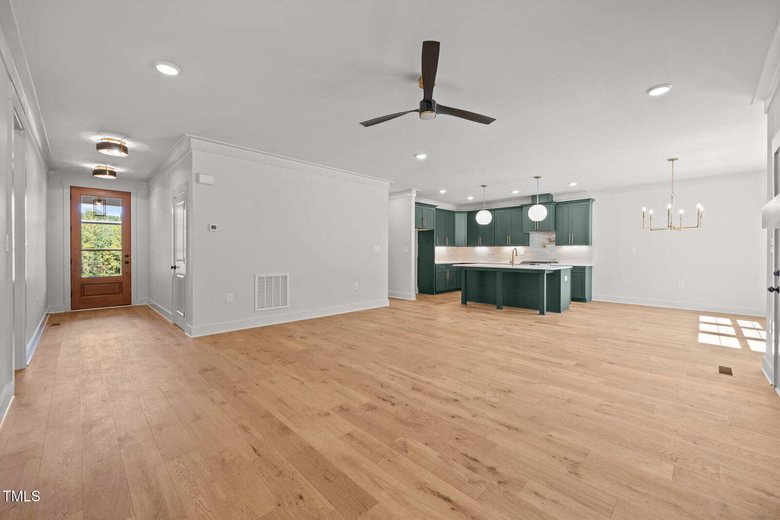Open-concept living space with light wood flooring, white walls, and a ceiling fan. The kitchen features green cabinets, white countertops, gold hardware, and modern light fixtures. A wooden front door is visible in the background.