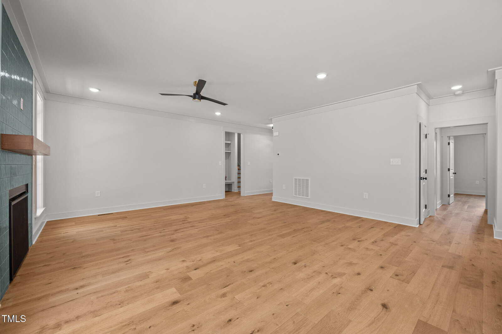 Spacious, modern living room with light wood floors, white walls, recessed lighting, a ceiling fan, a fireplace with a green tile surround, and an open doorway leading to other rooms.