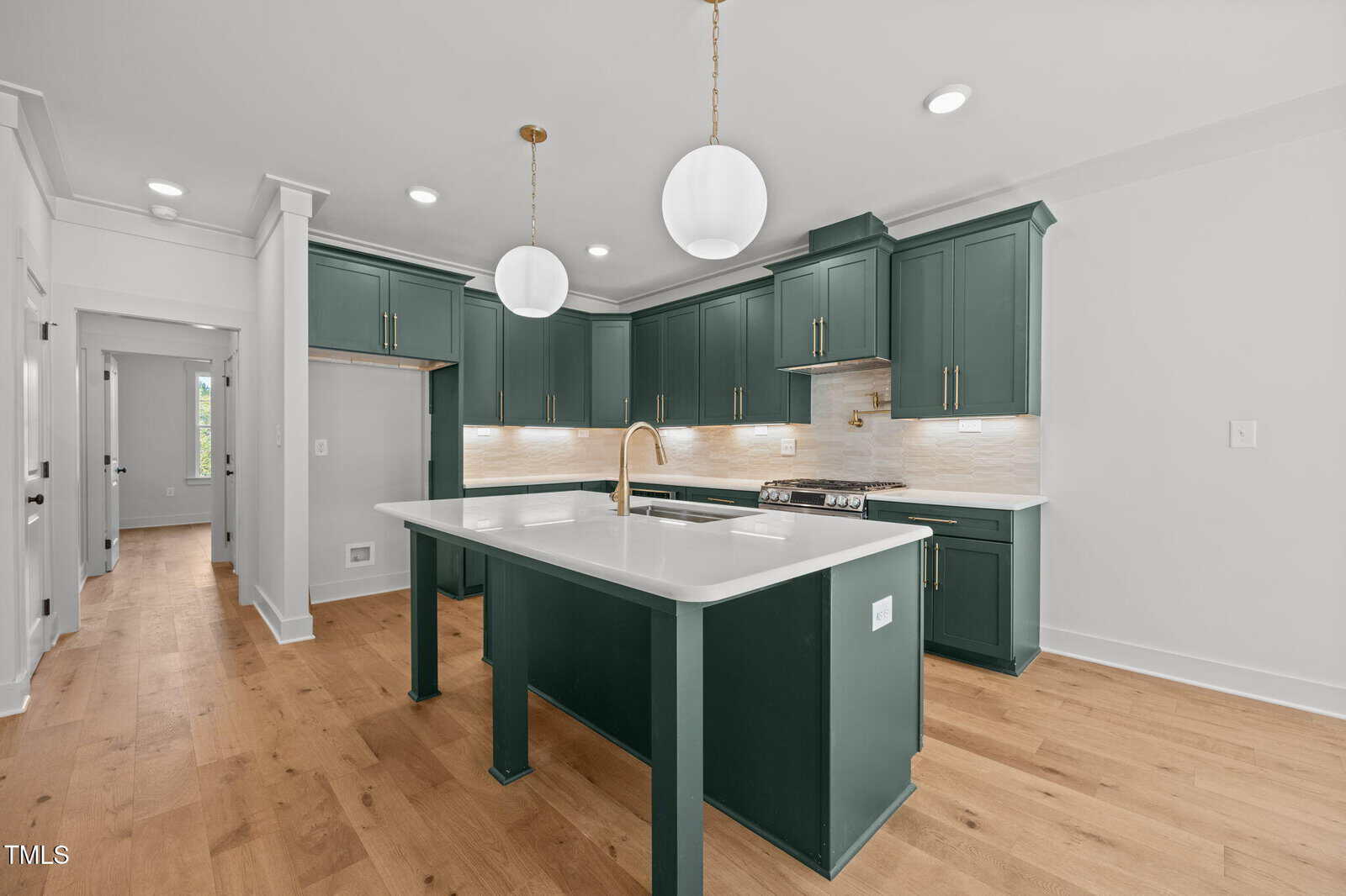 Modern kitchen with green cabinets, white countertops, light wood flooring, gold fixtures, two round pendant lights over the island, and stainless steel appliances. Light beige backsplash under cabinets.